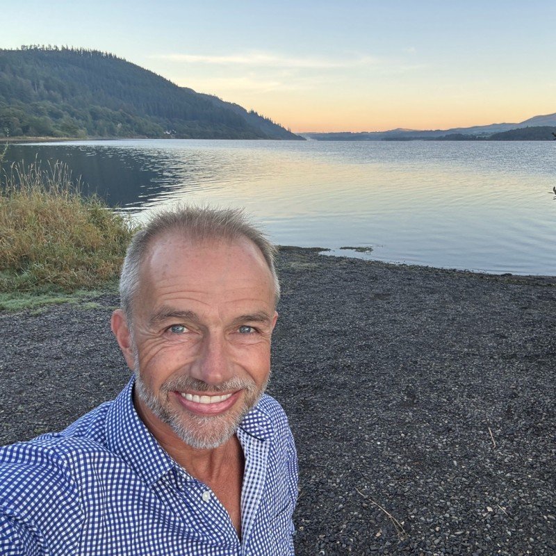 A smiling man with short gray hair and a beard taking a selfie near a lake during sunset, with hills in the background.