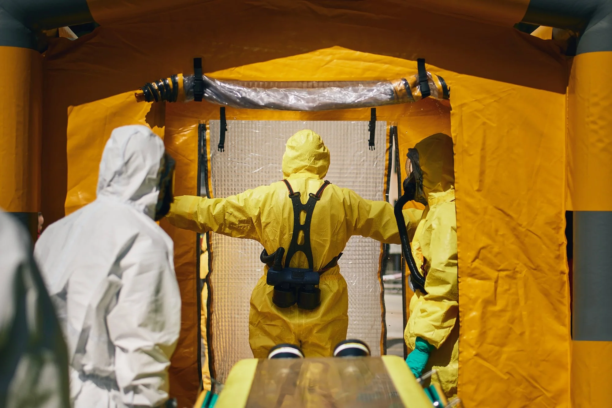 Medical personnel in yellow protective suits and masks entering a makeshift entrance with a yellow tunnel for a coronavirus or infectious disease quarantine or treatment area.
