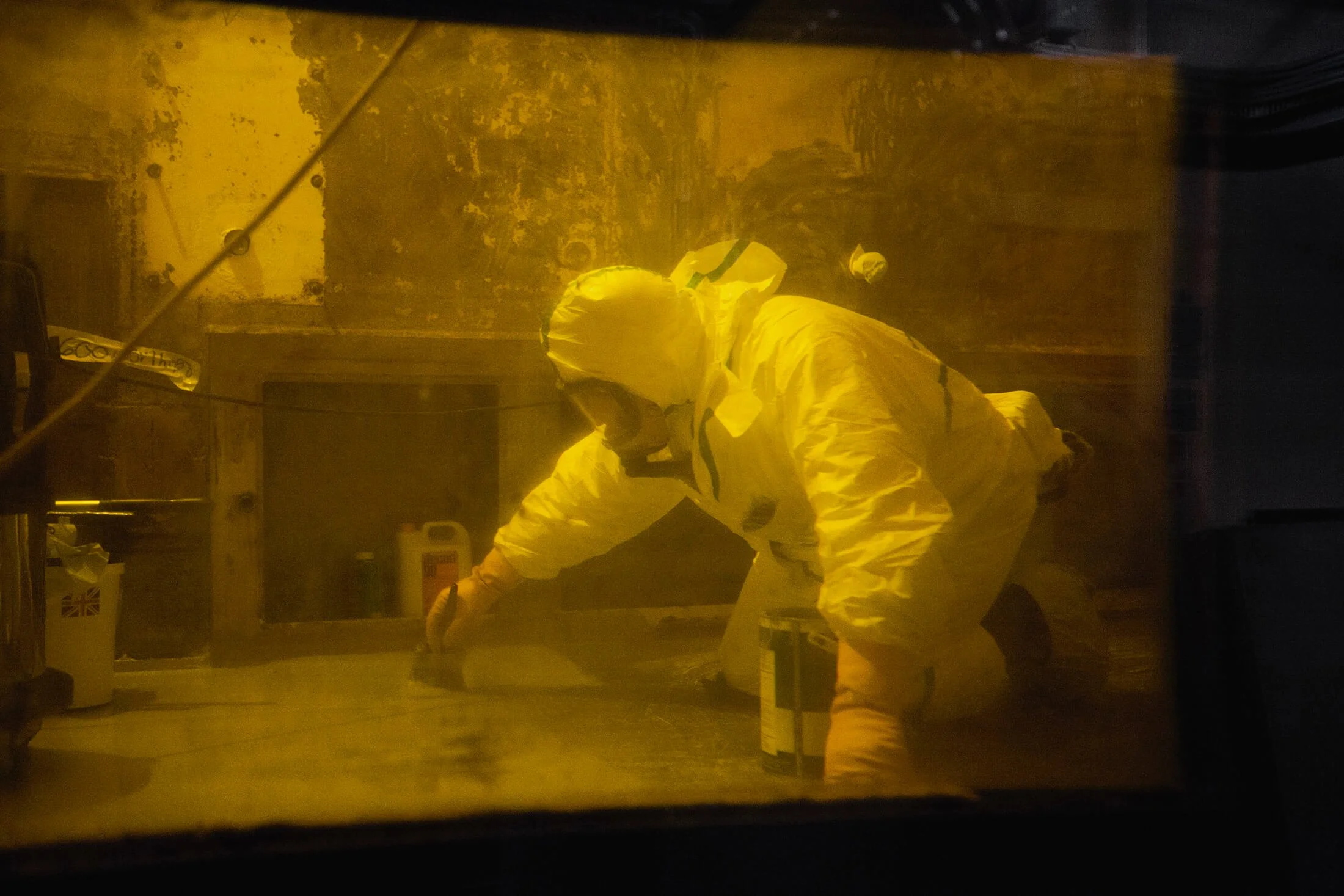 A person dressed in protective yellow hazmat suit working inside a room with a yellowish tint, seen through a window or glass partition.