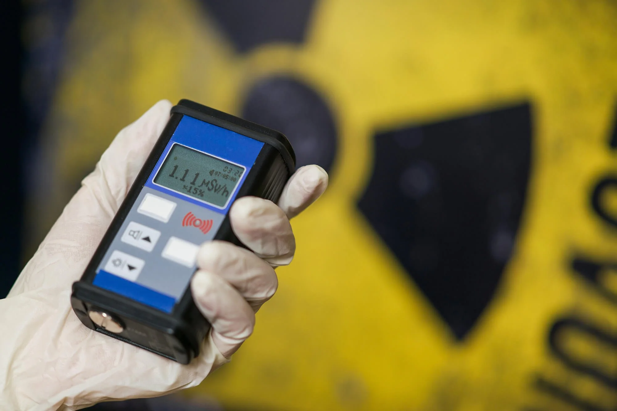 A person wearing a glove holds a handheld radiation detector measuring 1.11 microsieverts per hour, with a radiation hazard symbol in the background.