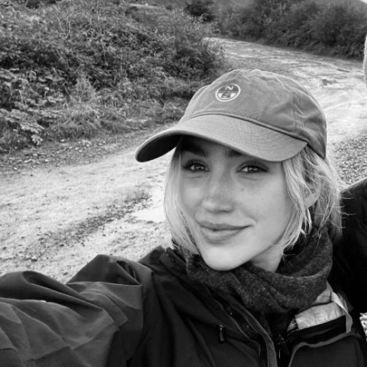 A young woman with blonde hair, wearing a baseball cap and a jacket, taking a selfie outdoors on a trail with trees and bushes in the background.