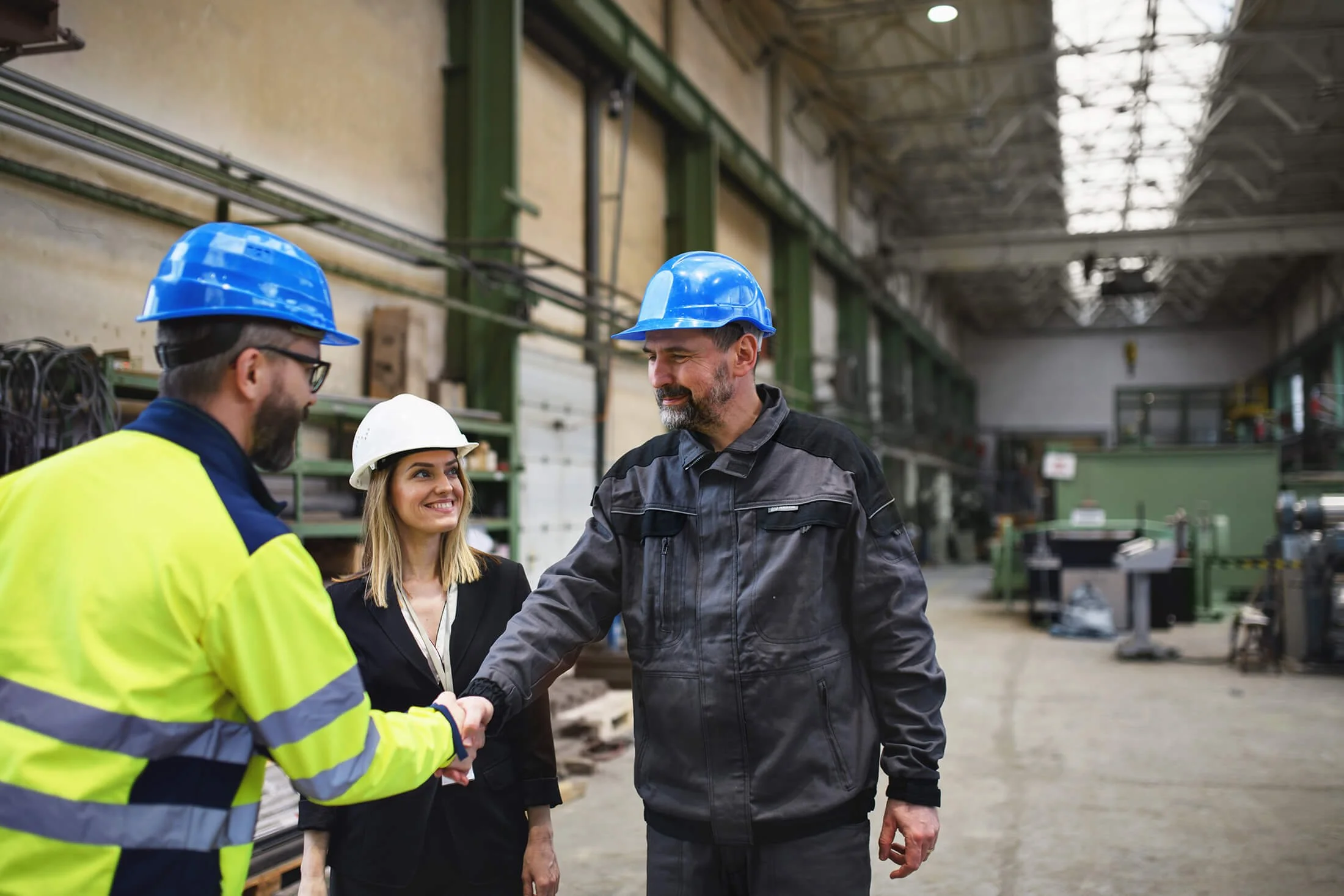 Three people wearing safety helmets shaking hands inside a large industrial warehouse, with machinery and equipment in the background.