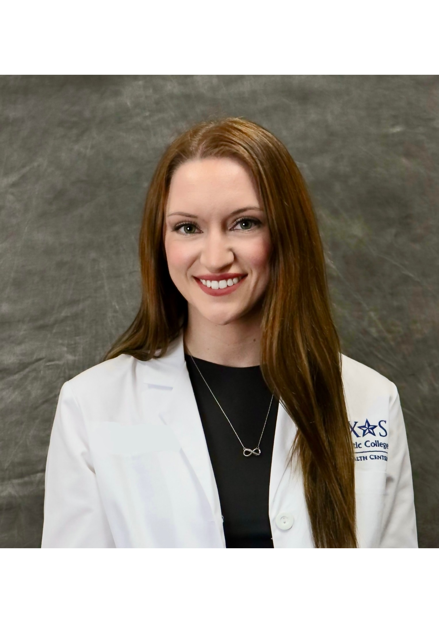 Smiling woman with long red hair wearing a white lab coat and a necklace with an infinity symbol, standing against a gray textured background.