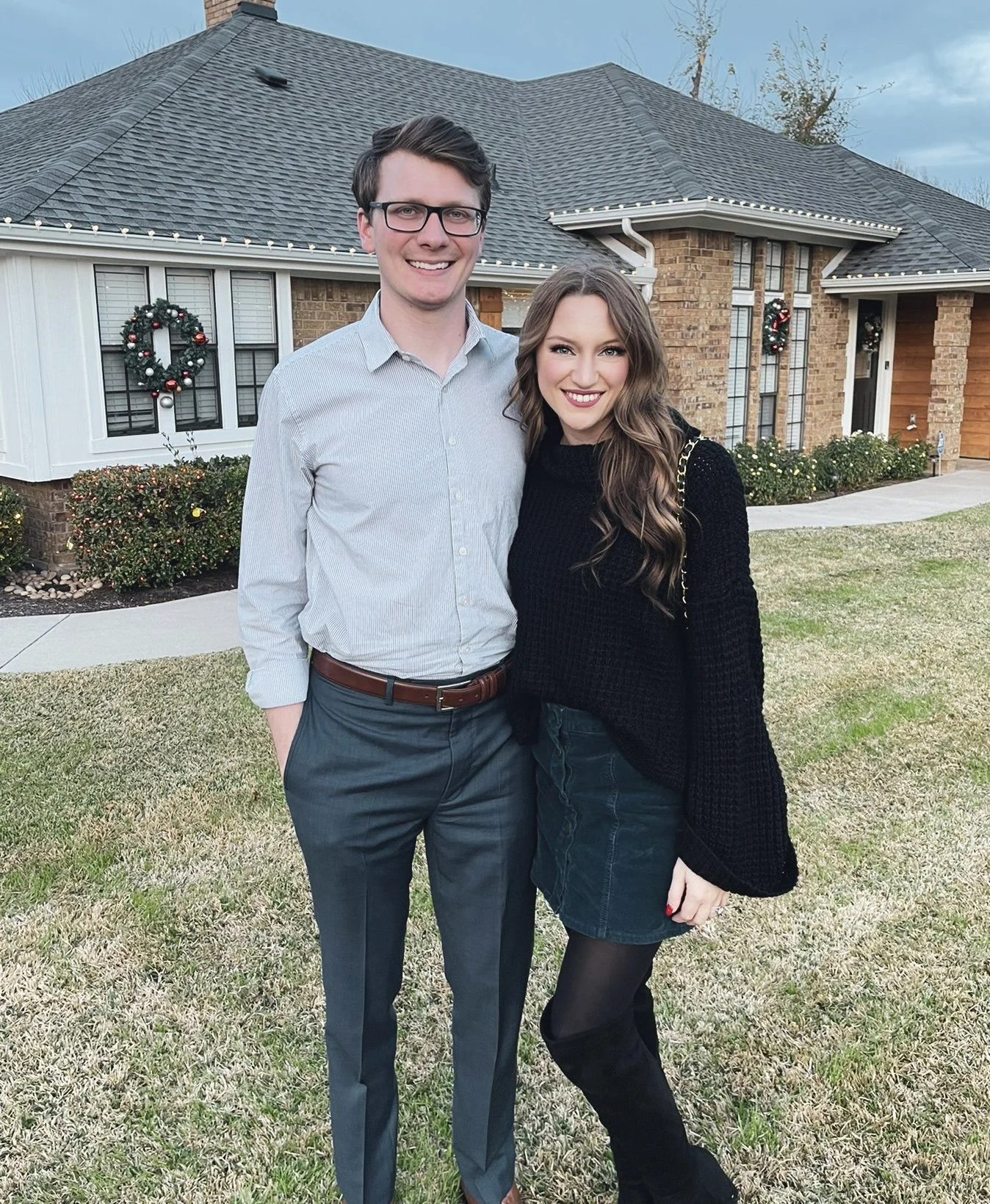 A young man and woman standing outdoors in front of a brick house decorated with Christmas wreaths, smiling at the camera.
