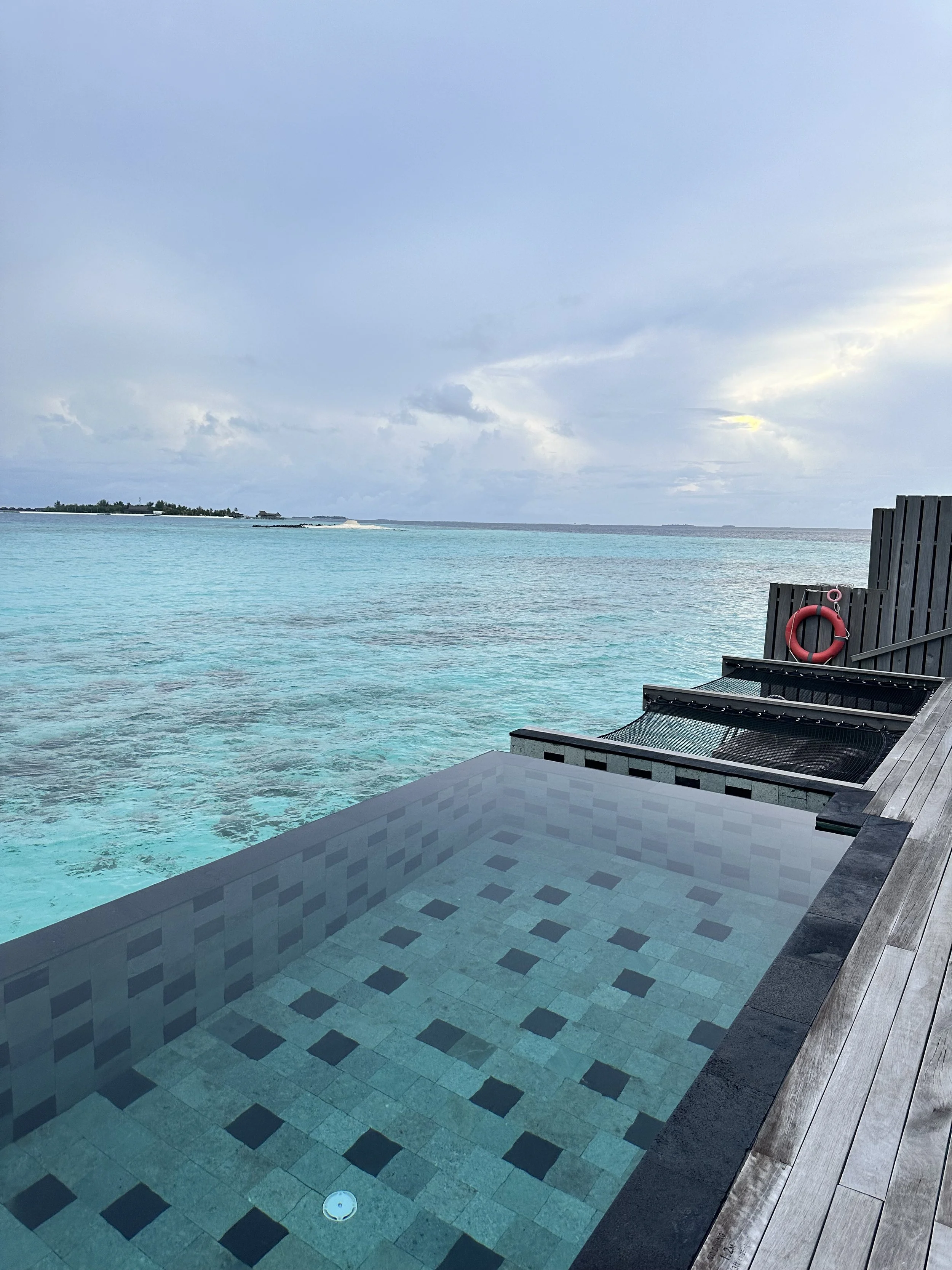 Overwater infinity pool overlooking the ocean with clear turquoise water, cloudy sky in the background, and a fire rescue ring on a wooden privacy wall.