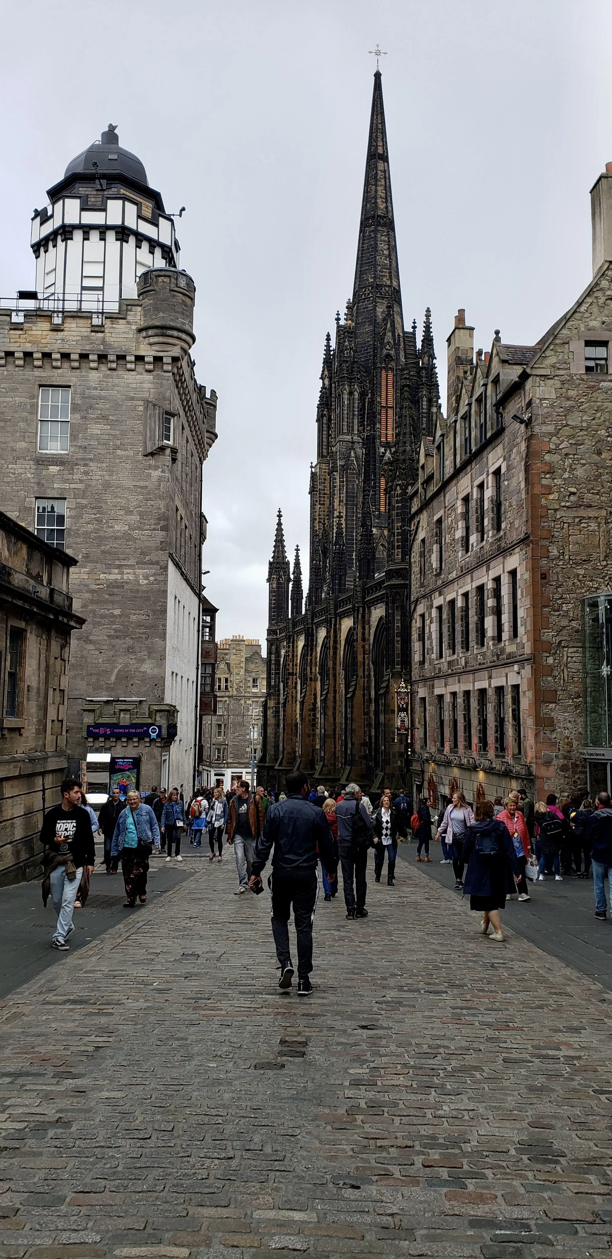 A bustling street scene with a Gothic-style church with a tall spire and intricate architecture, surrounded by historic stone buildings and a crowd of people walking on cobblestone pavement.