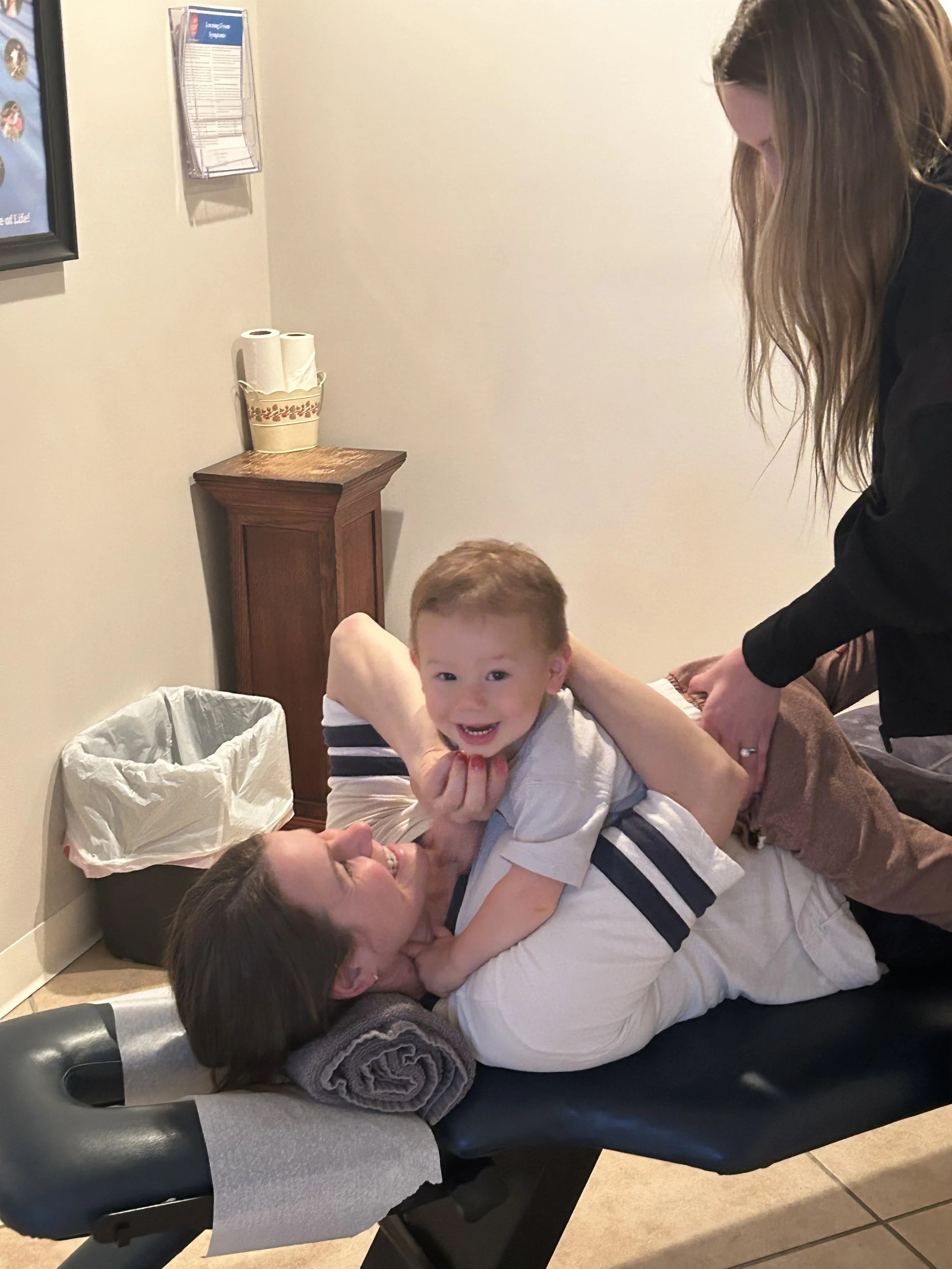 The child happily smiles while getting an adjustment. The child is lying down on his mother, who is laughing.