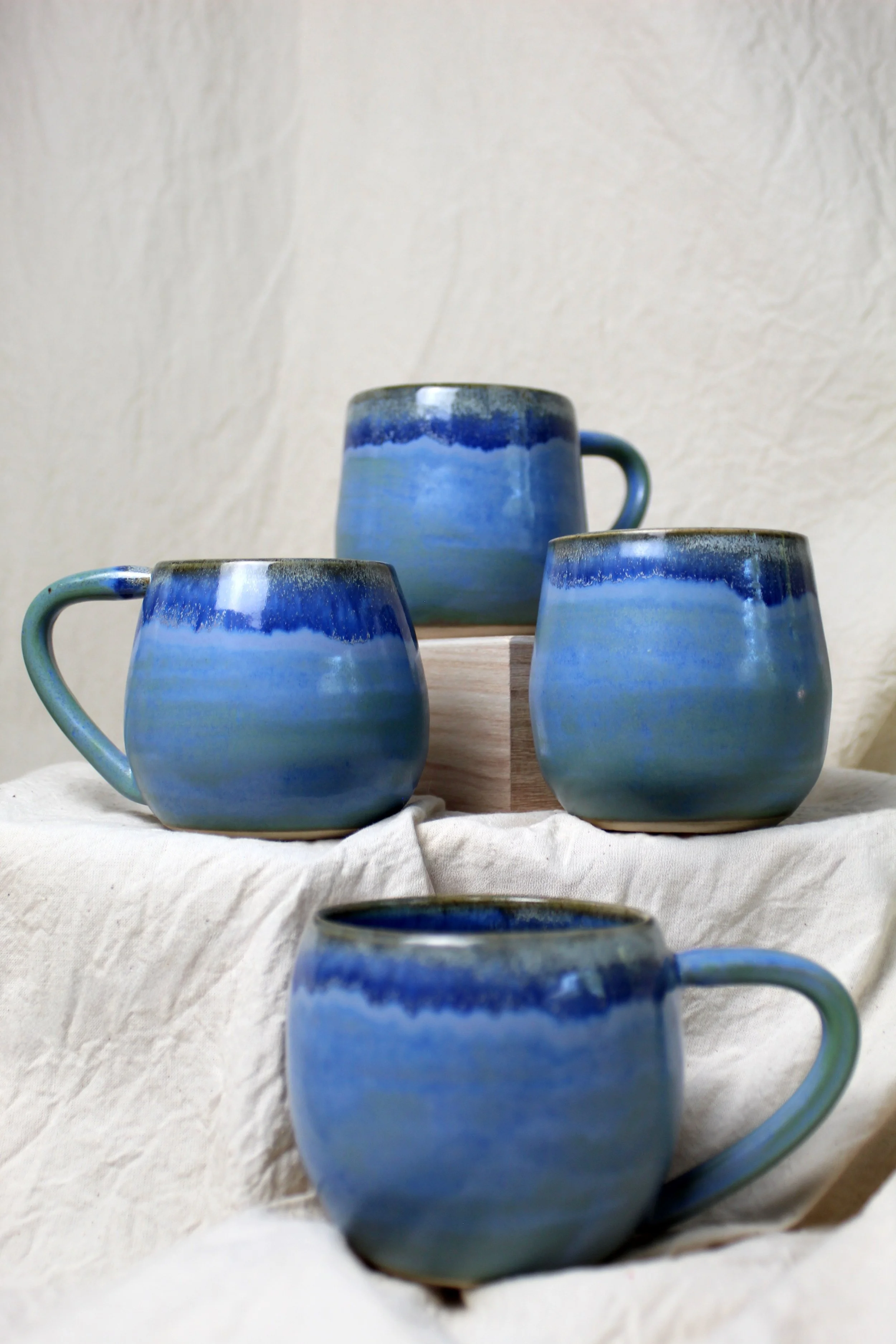 Four ceramic coffee mugs with blue and green glazed patterns arranged on a fabric surface with a neutral textured background.