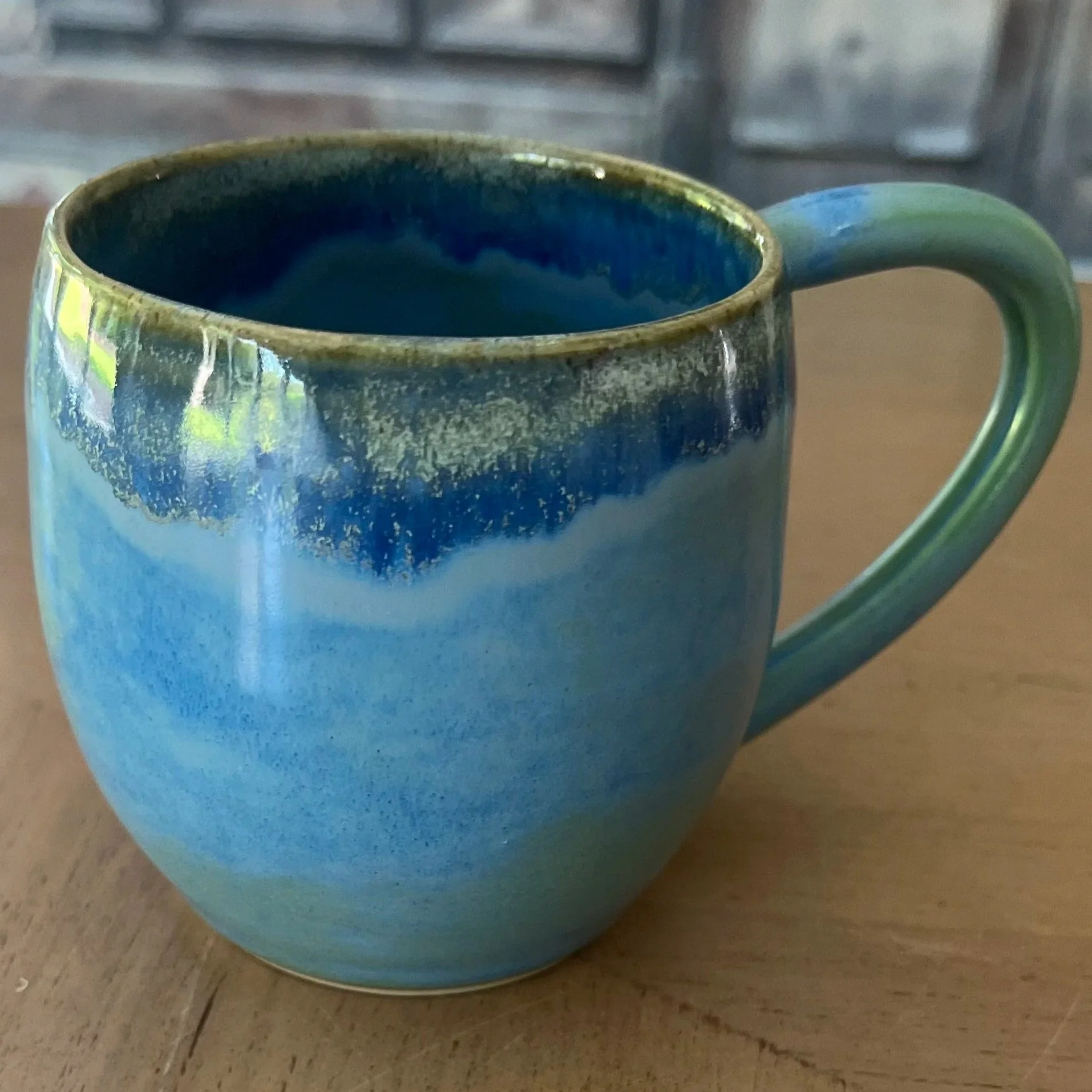 Handmade stoneware mug with a layered glossy glaze in shifting tones of sky blue, deep ocean blue and mint green, sitting on a wooden surface.