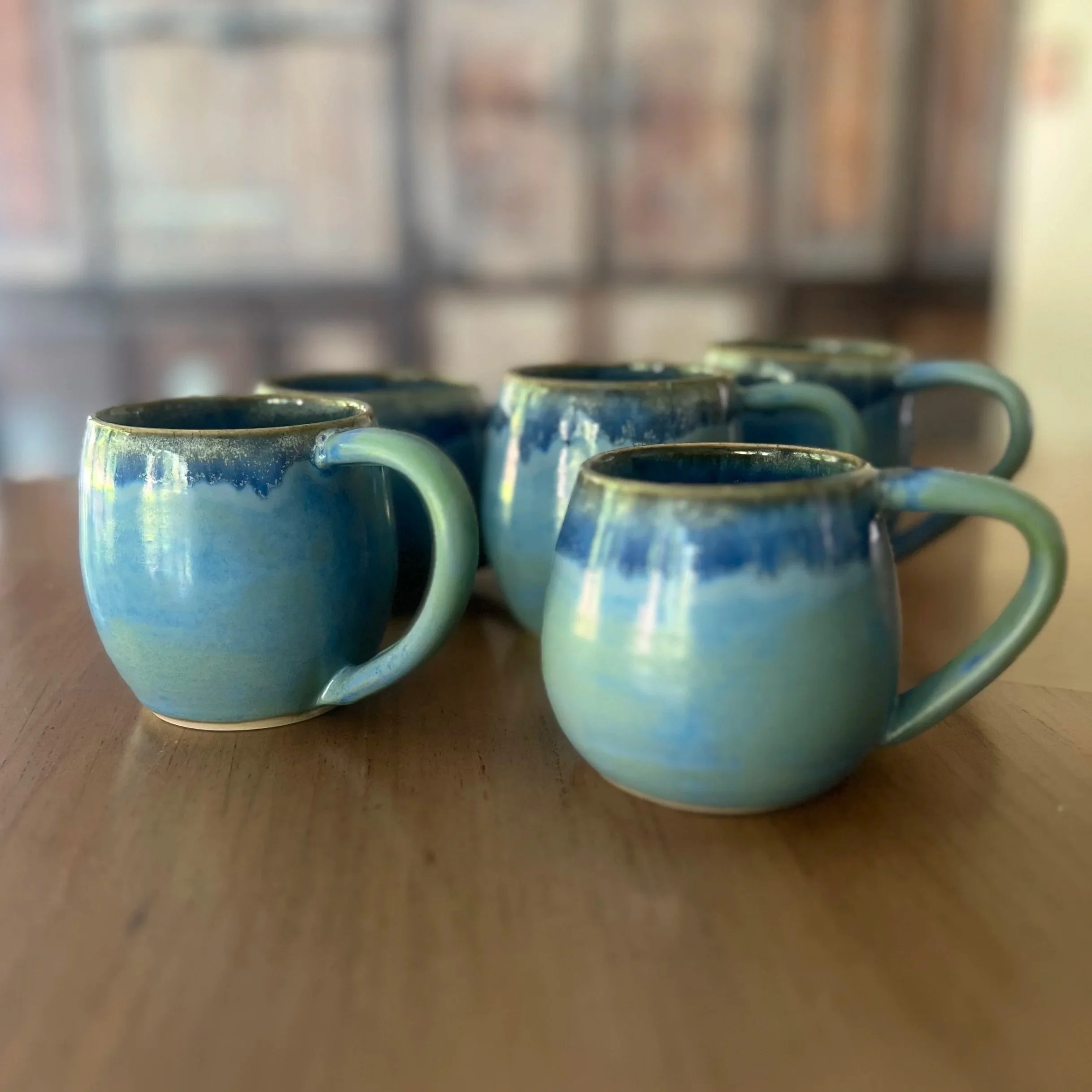 Five handmade stoneware mugs with a layered glossy glaze in shifting tones of sky blue, deep ocean blue and mint green, sitting on a wooden surface.