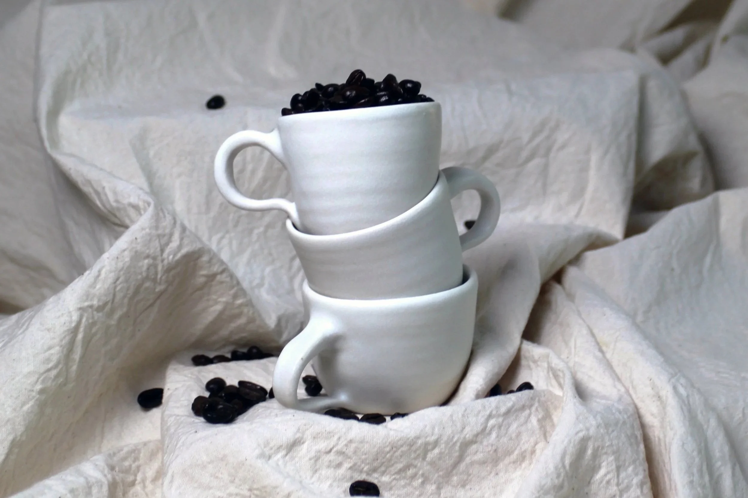Three stacked white ceramic coffee cups with black coffee beans inside the top cup, placed on crumpled white fabric backdrop.