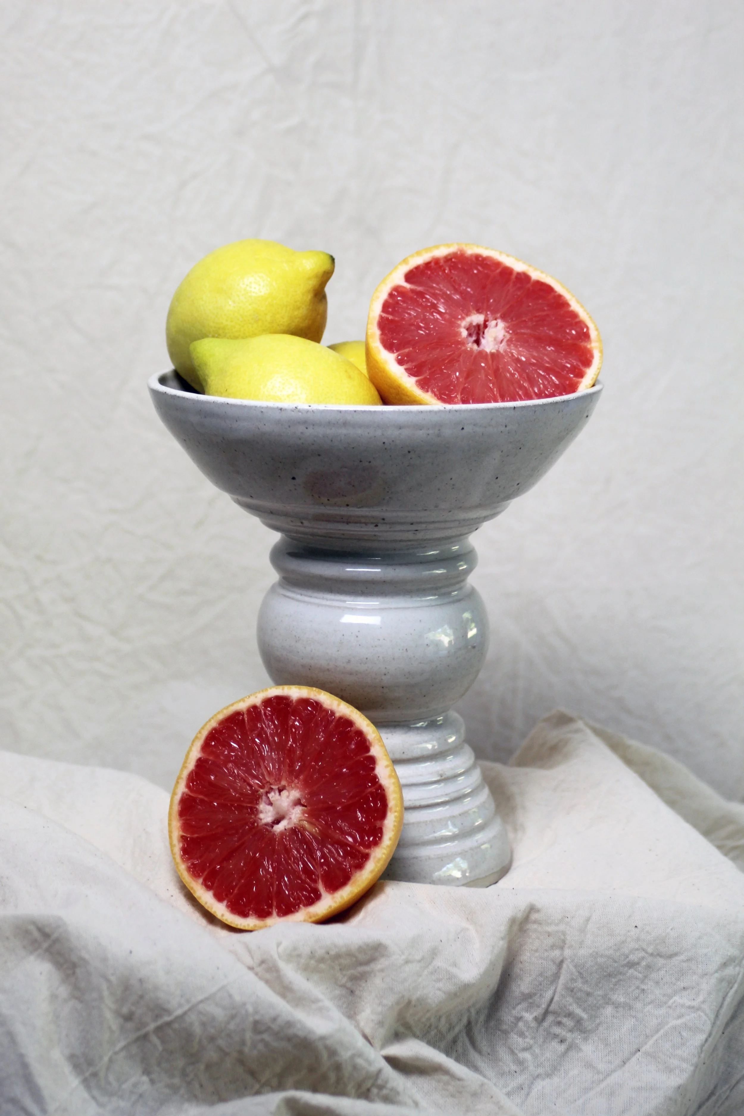 A white ceramic pedestal bowl containing two lemons and two halves of a pink grapefruit, with one grapefruit half resting on a white cloth.