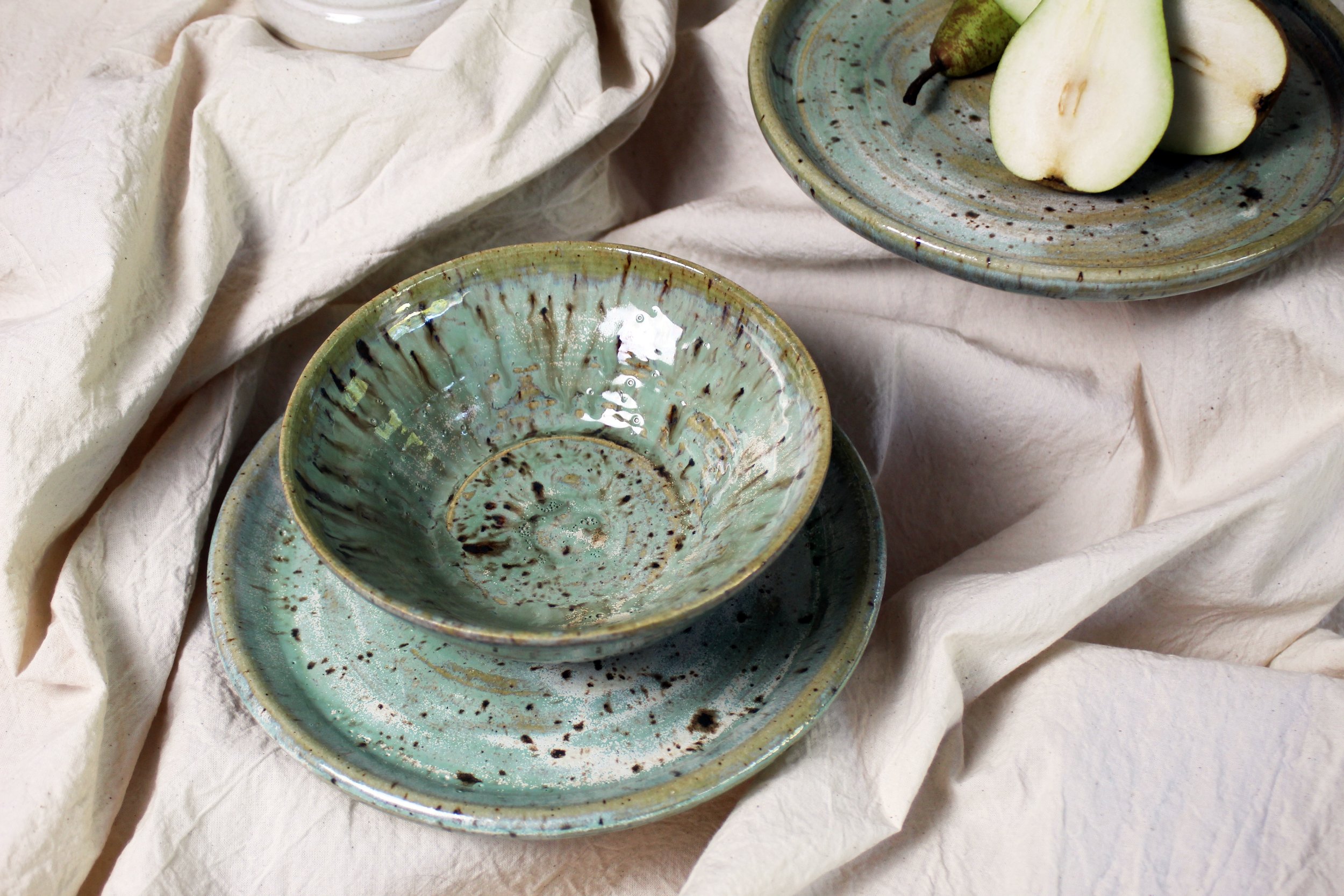 A set of three ceramic bowls and plates with a splattered green glaze finish, one bowl stacked on a plate, with pears and halved pears on a plate, all placed on cream-colored fabric.