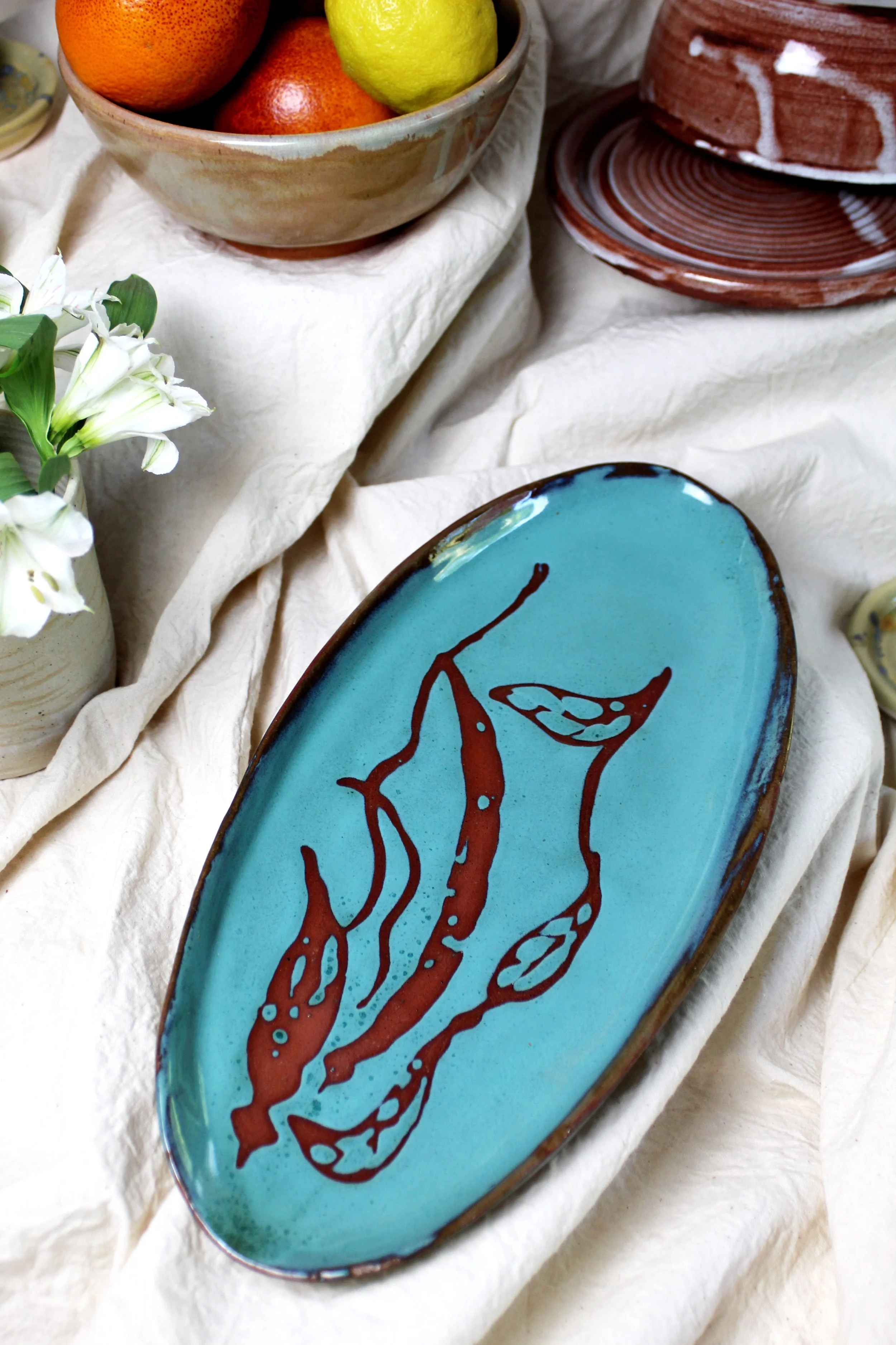 A decorative ceramic plate with abstract red-brown design on a turquoise background, placed on crumpled off-white fabric, alongside a vase with white flowers, a bowl with oranges and lemons, and a stack of red and white striped plates.