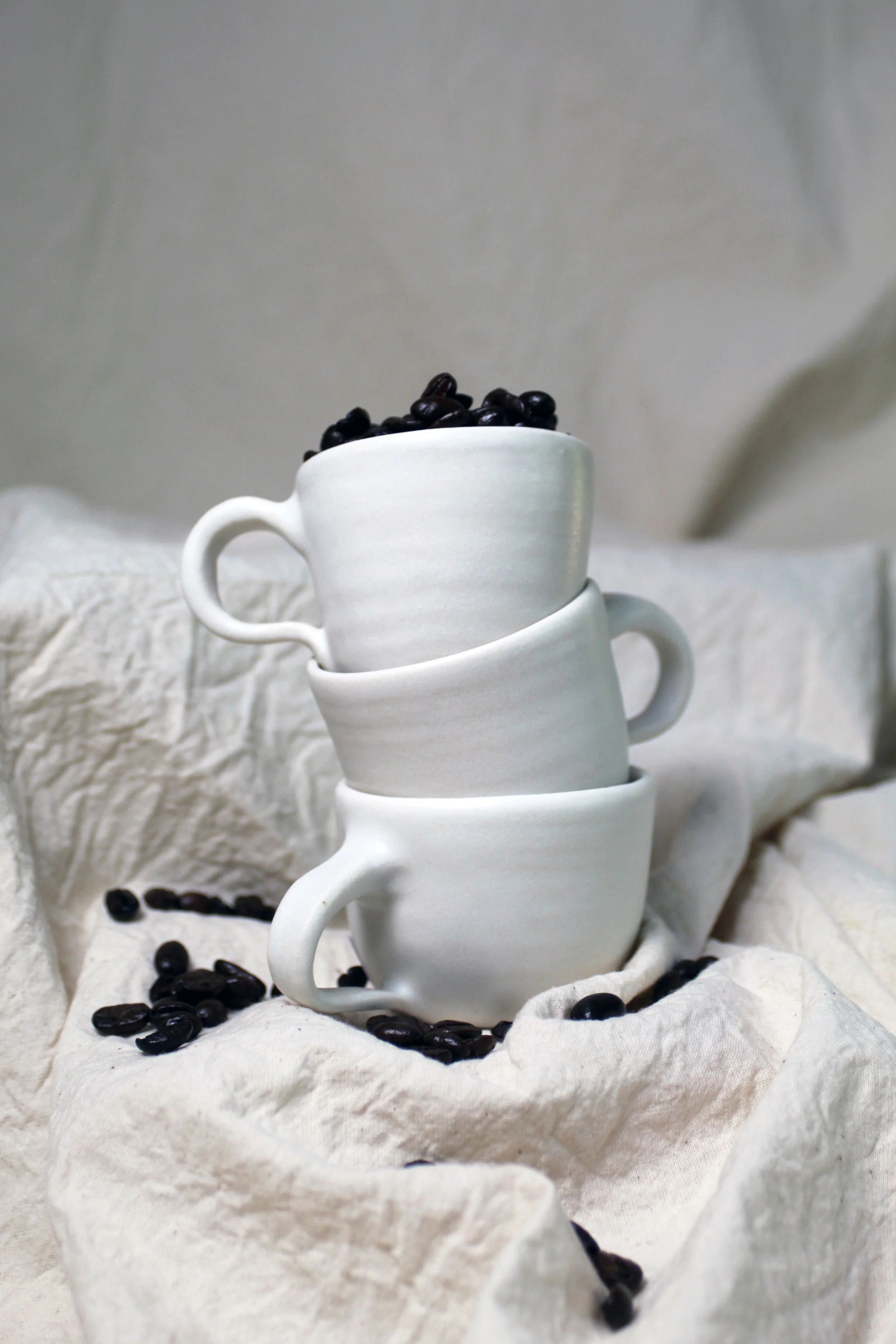 Three white ceramic cups stacked with black coffee beans on top, with some beans spilled around on a soft, light-colored fabric surface.