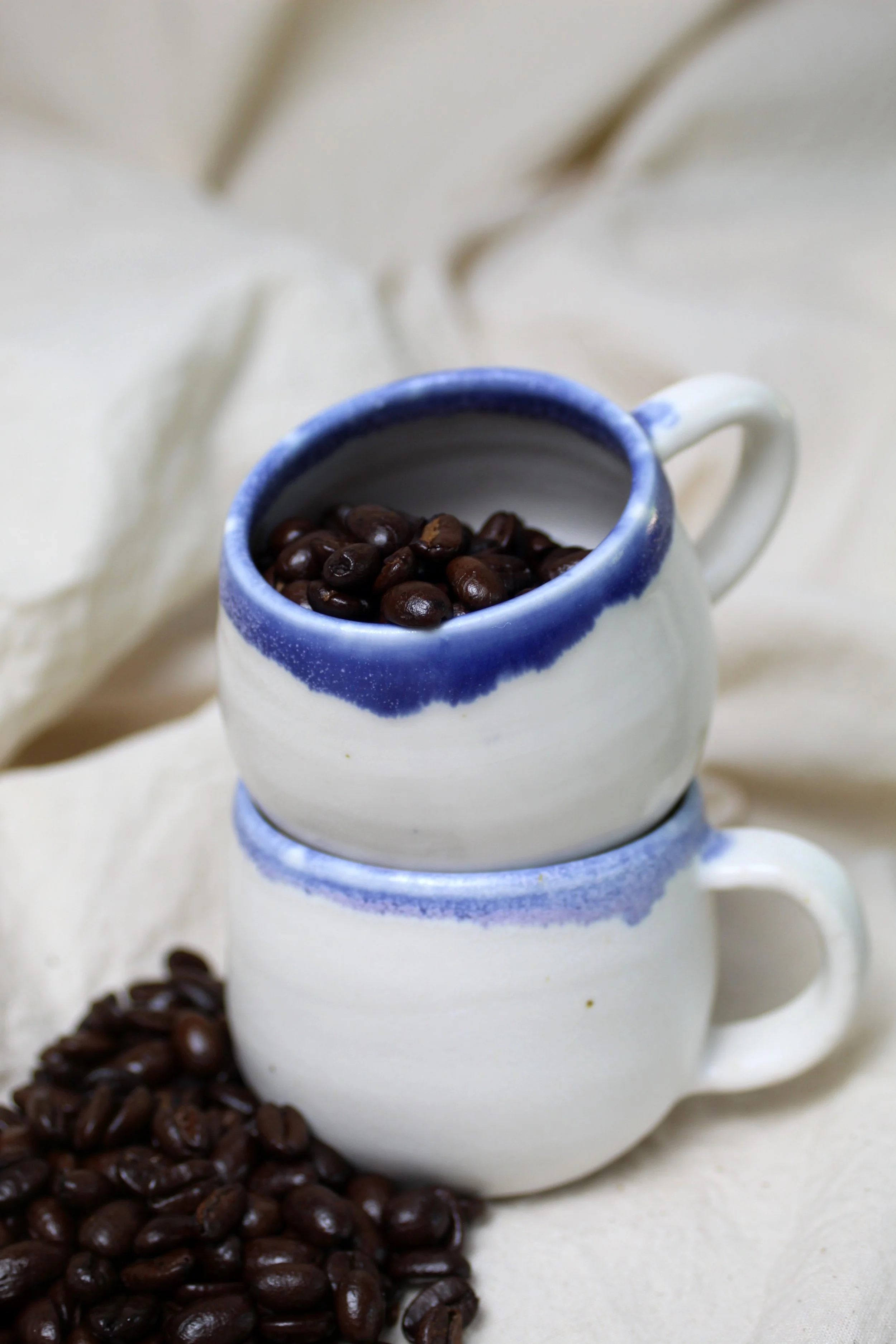 Two small white ceramic mugs with blue rims, one stacked on top of the other. The top mug is filled with dark roasted coffee beans. Some coffee beans are also spilled on the surface in front of the mugs, with a soft, cream-colored fabric in the backg