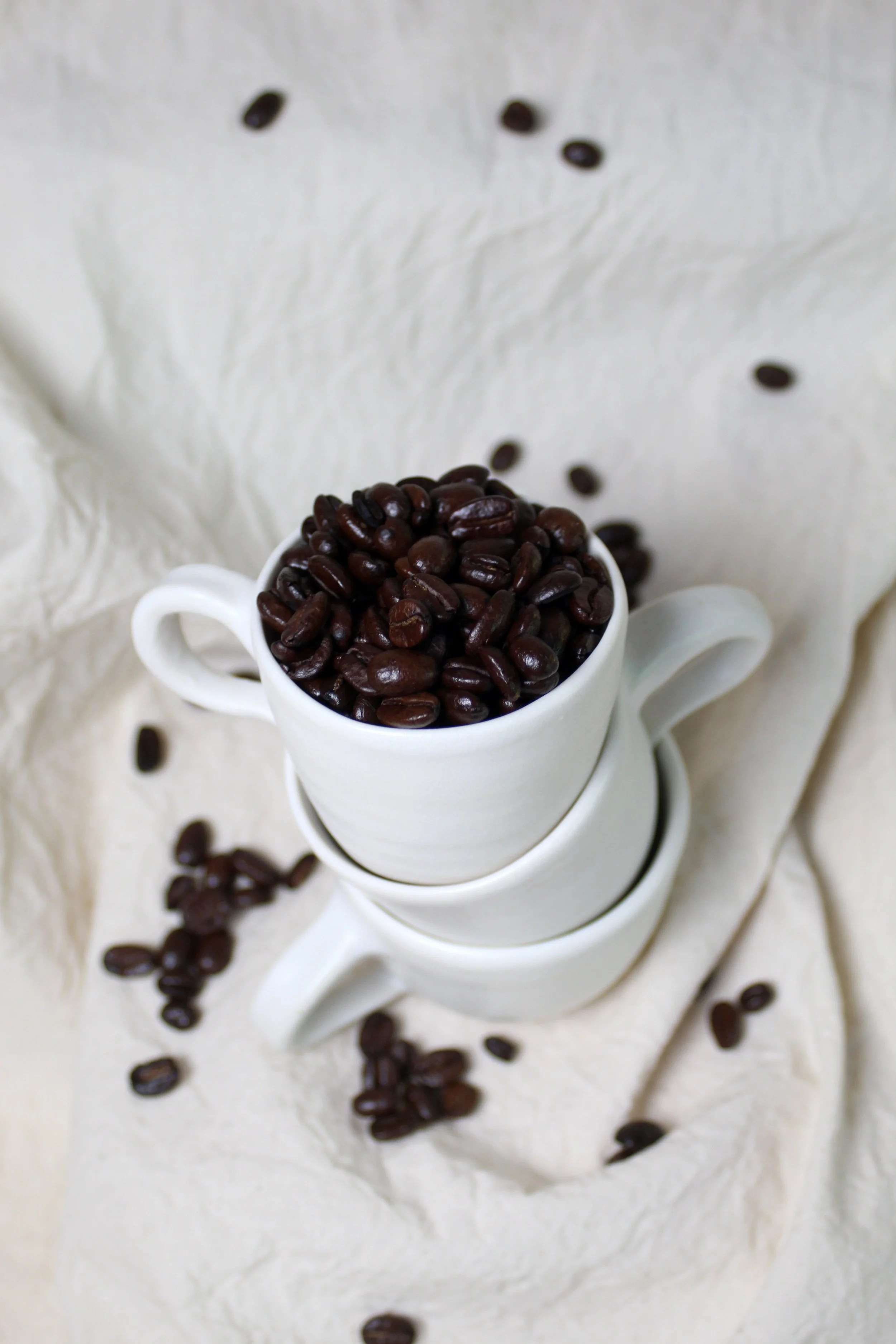 A white ceramic cup filled with dark roasted coffee beans, placed on matching cups, with scattered beans on a cream-colored cloth background.