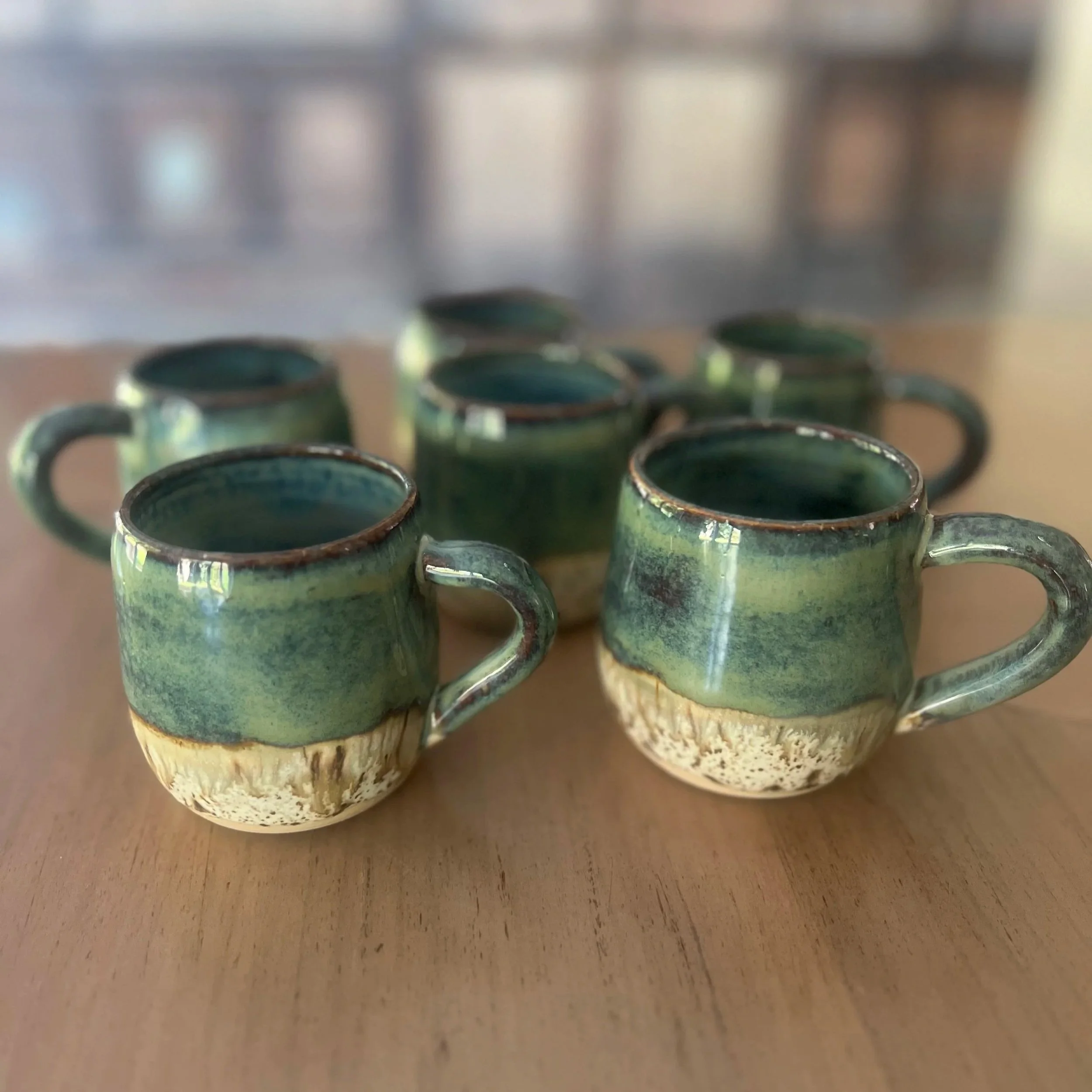 Six handmade stoneware teacups with a glossy forest green glaze fading into a textured foamy cream base, sitting on a wooden surface.