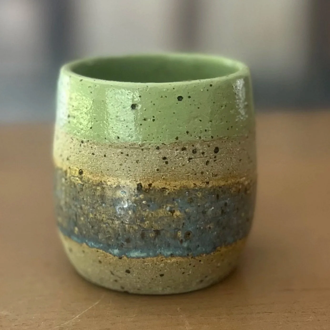 Handmade stoneware tumbler cup with a glossy sage green rim and layered sandy, ochre and grey speckled clay body, sitting on a wooden surface.