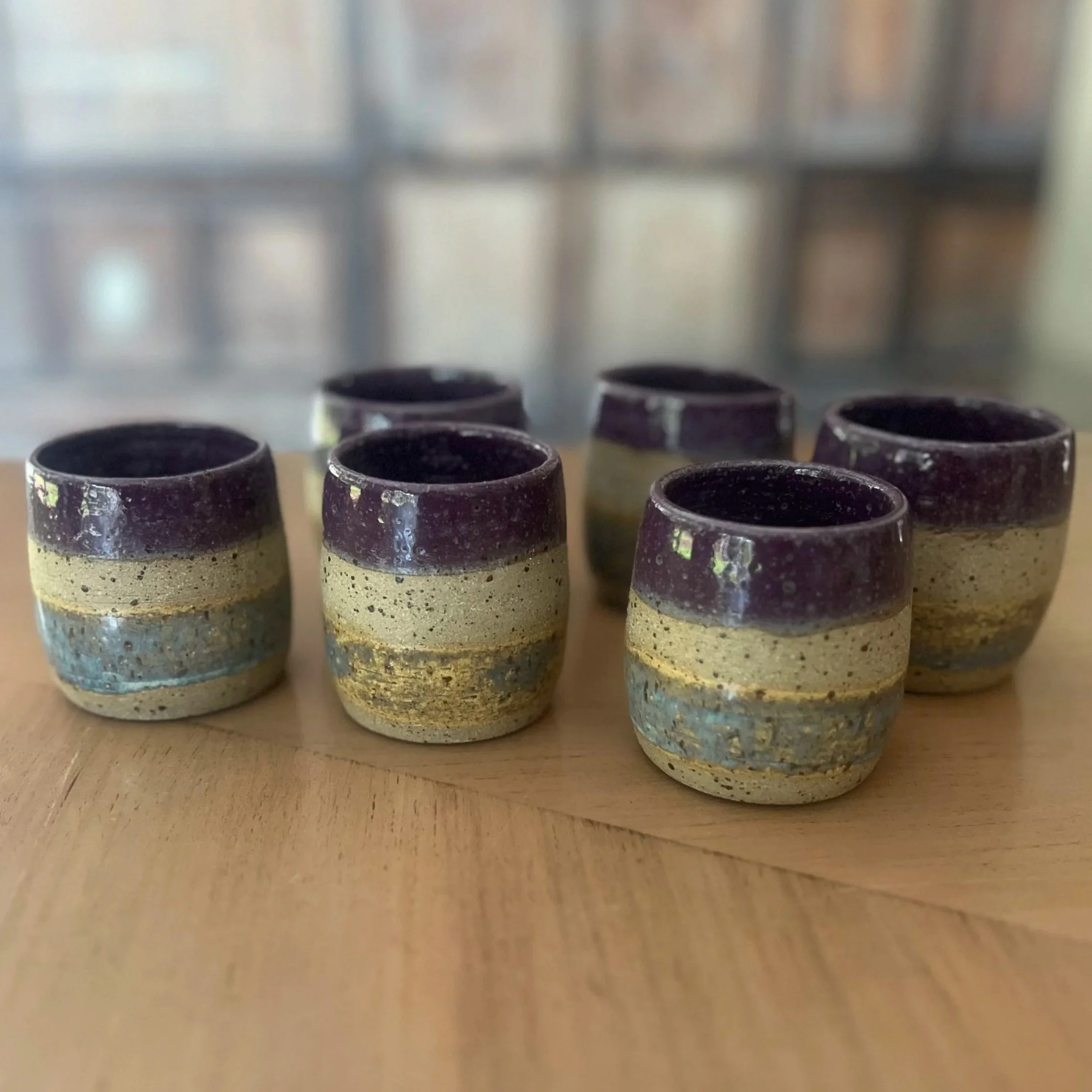 12:59Six handmade stoneware tumbler cups with varying glossy aubergine coloured rims and layered speckled clay bodies, arranged together on a wooden surface.