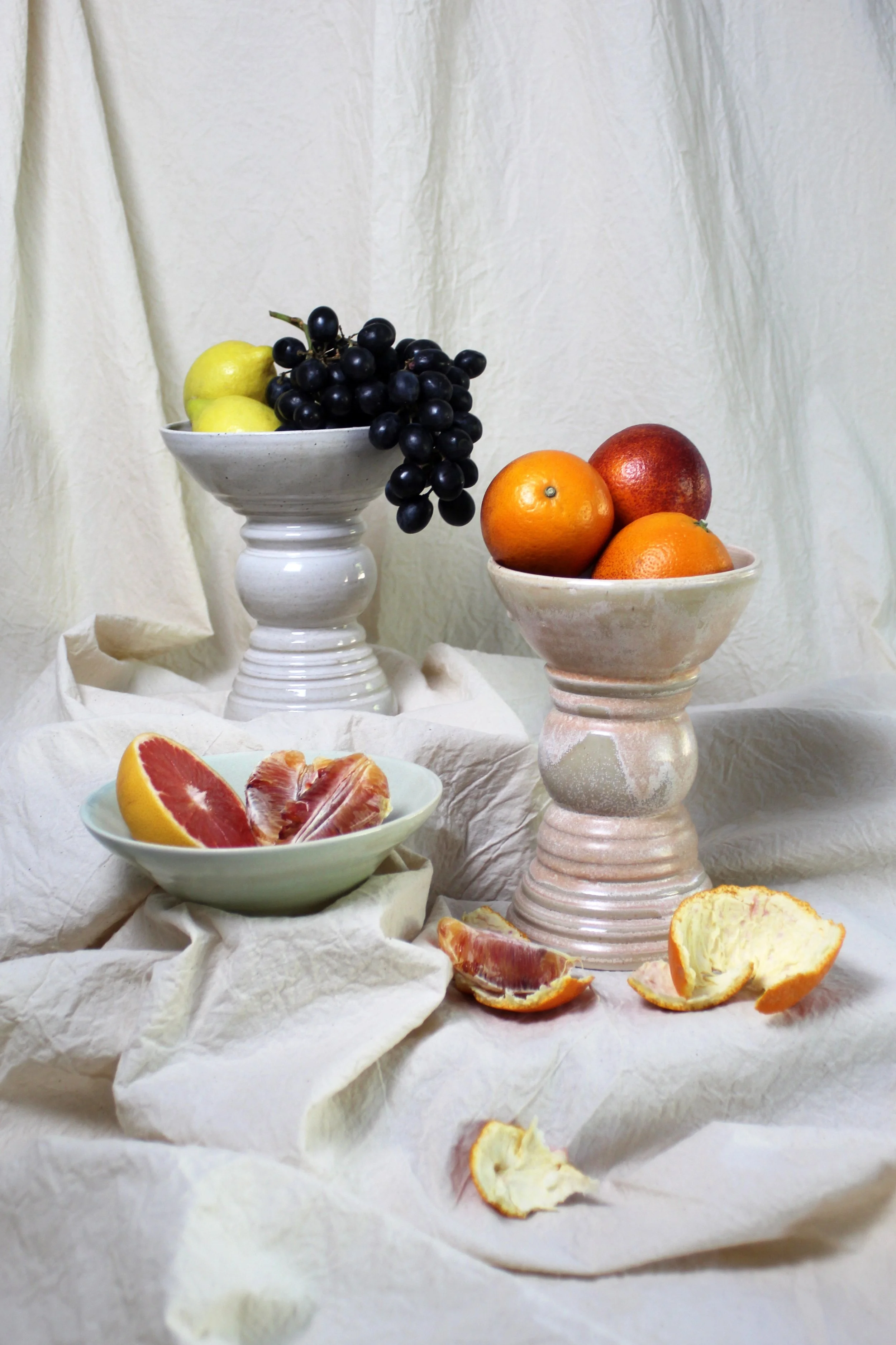 Assorted fresh fruits including grapes, oranges, grapefruit, apples, and apple peels arranged on vintage-style stands and a cloth background.