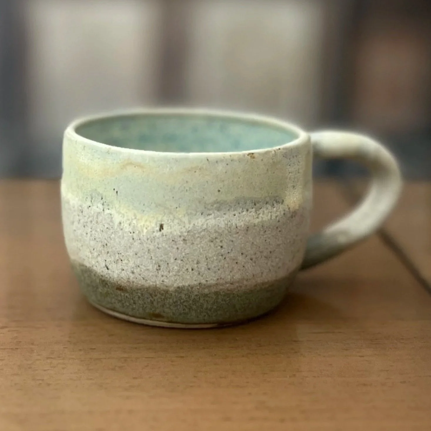 Handmade stoneware mug with a soft matte white and sandy speckled exterior and a pale celadon blue interior glaze, sitting on a wooden surface.