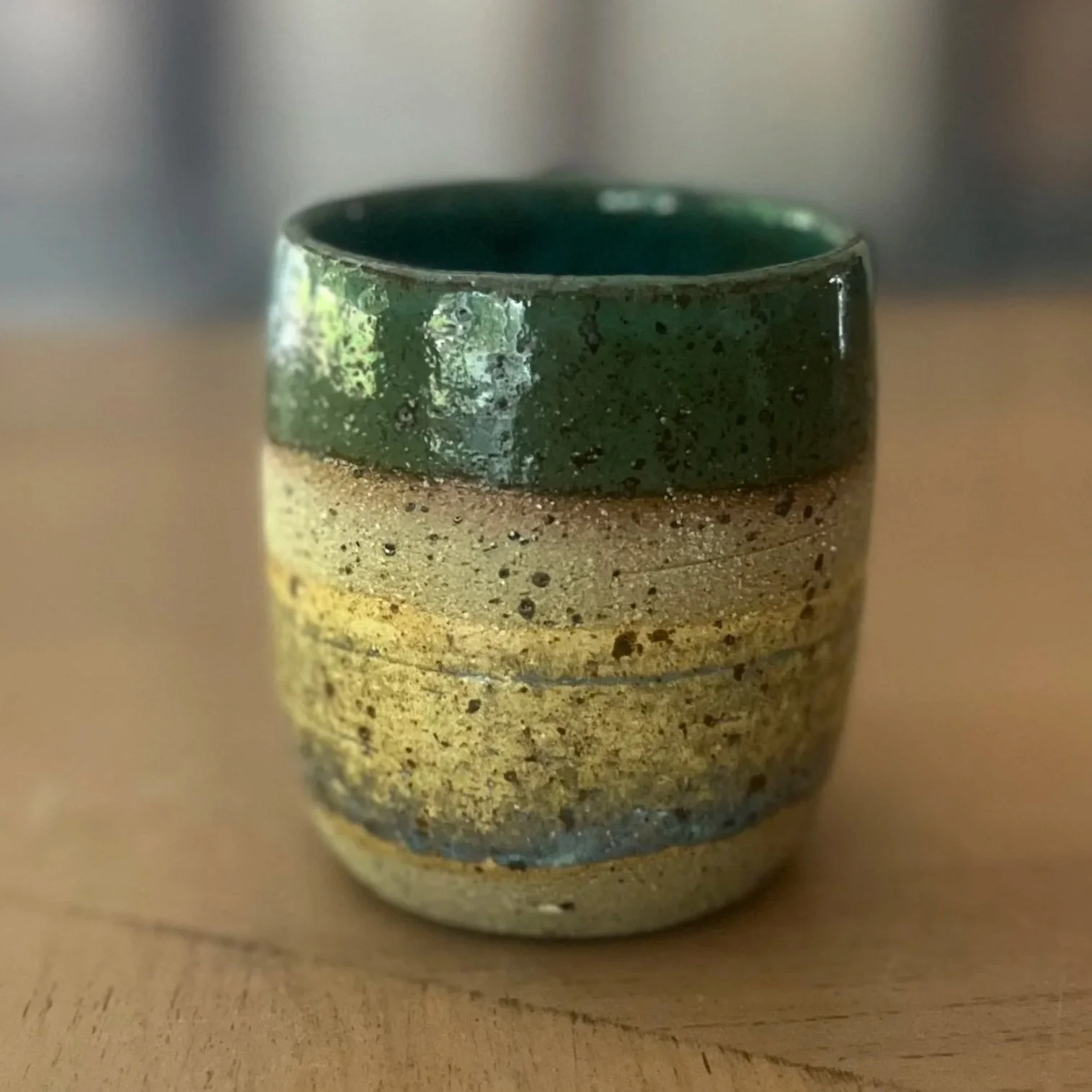 Handmade stoneware tumbler cup with a glossy deep green rim and layered sandy, ochre and grey speckled clay body, sitting on a wooden surface.
