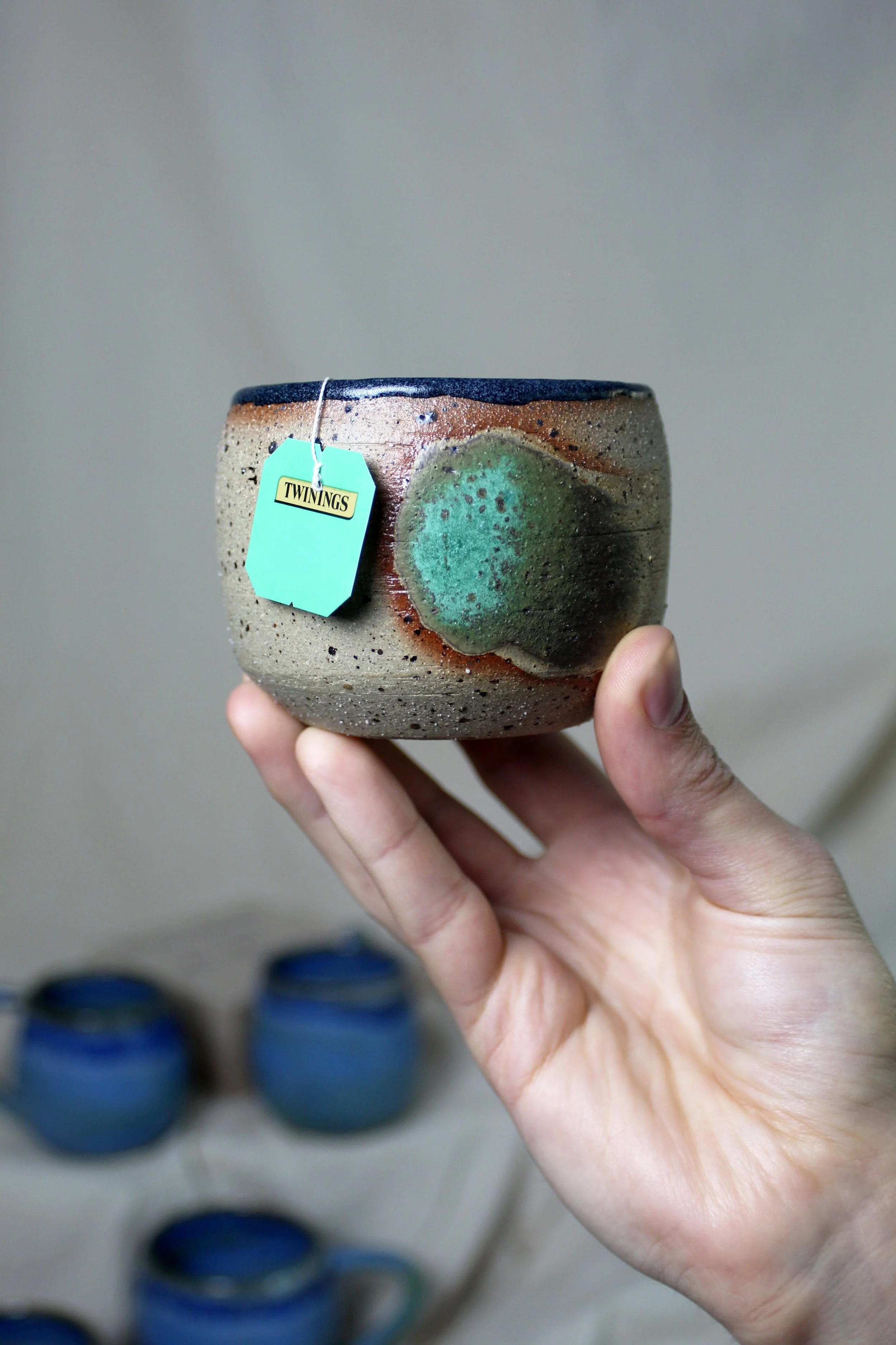 Hand holding a speckled ceramic bowl with a green glaze design, tagged with a Twinings label, with blue cups blurred in the background.
