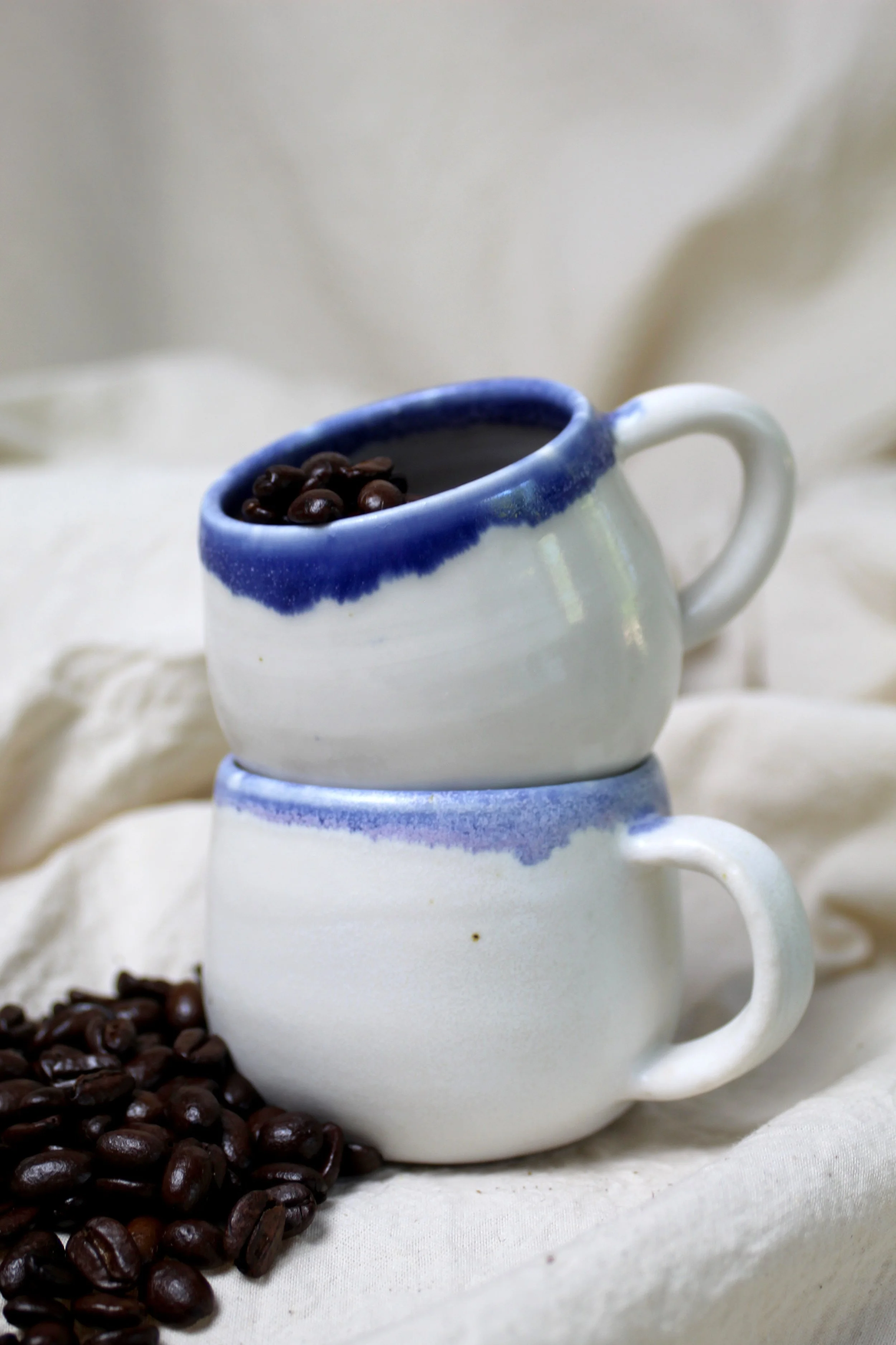 Two white ceramic coffee mugs with blue accents, stacked on top of each other, filled with coffee beans and placed on a soft, light-colored fabric.