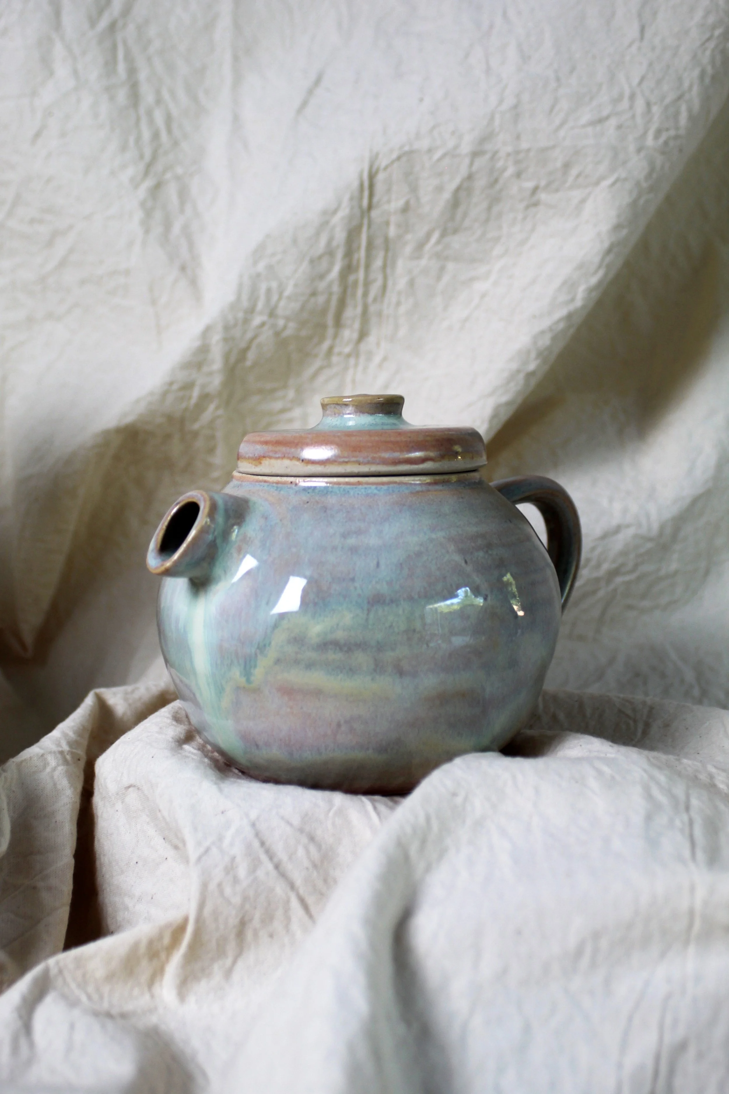 Colorful ceramic teapot with a lid, spout, and handle, resting on a light-colored fabric with a textured cream background.
