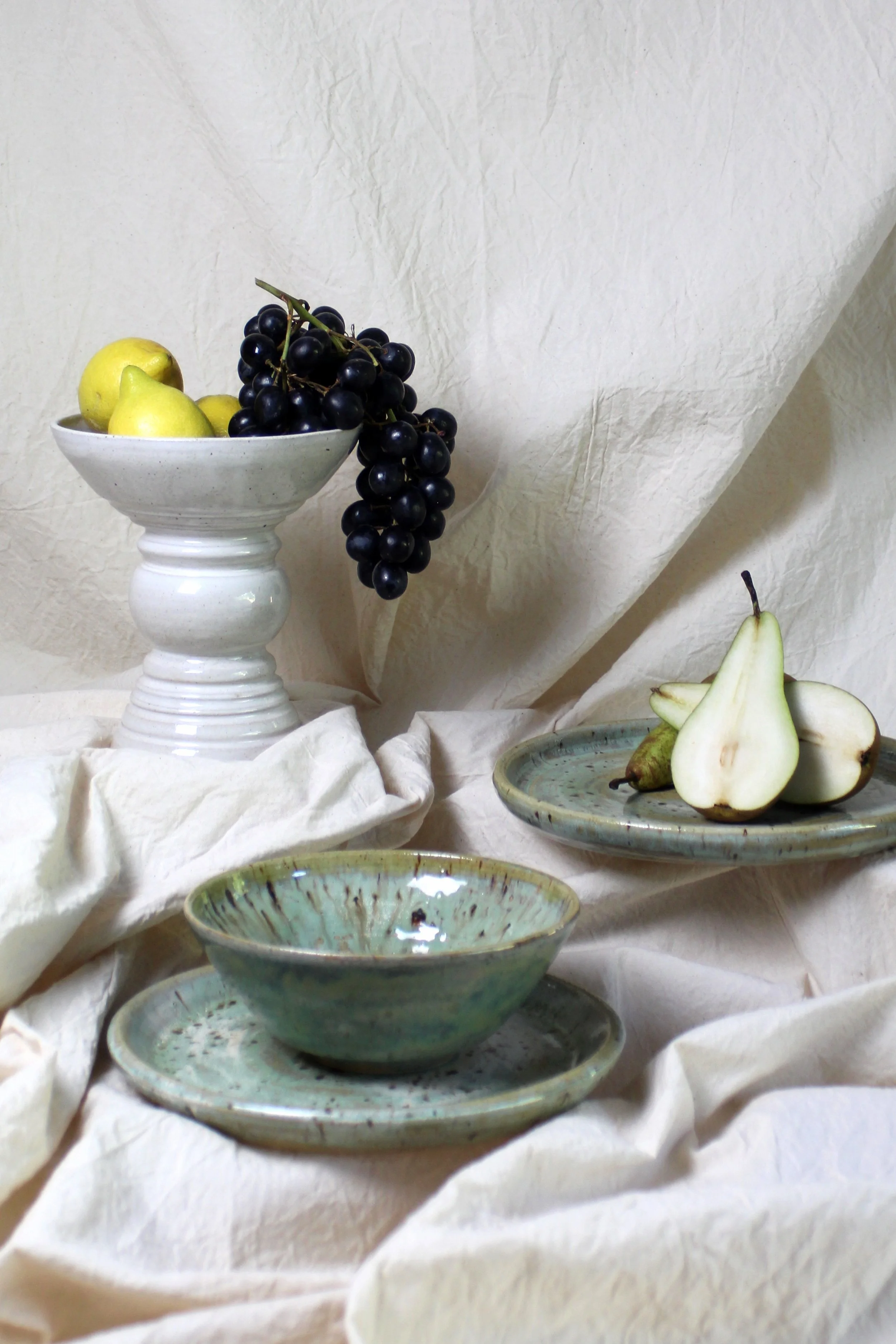Still life of black grapes, yellow pears, and green pears on ceramic plates and a pedestal bowl, with cream-colored cloth backdrop and table covering.