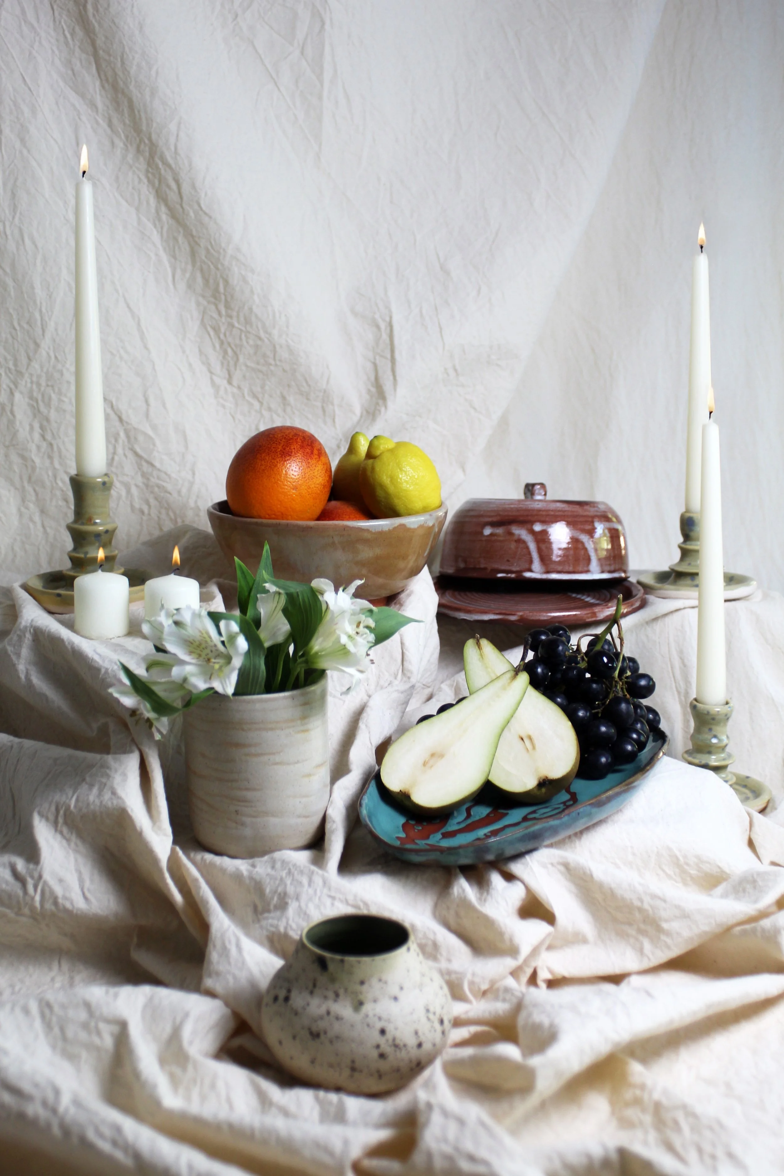 Still life of fruit and flowers on a crinkled fabric backdrop with candles and ceramic dishes.