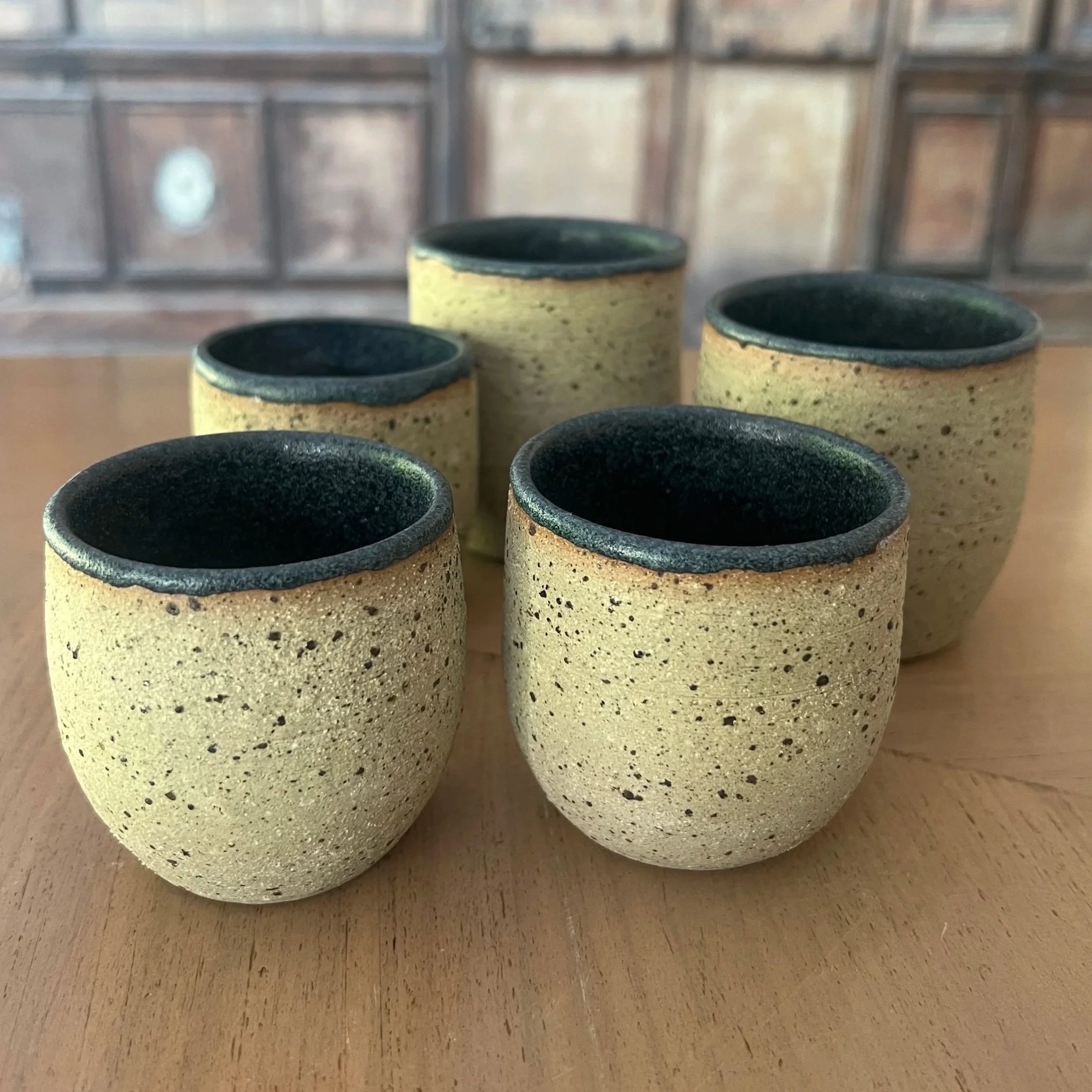 Five handmade stoneware tumbler cups with a speckled sandy yellow lava-flecked exterior and deep black granite rim and interior, sitting on a wooden surface.