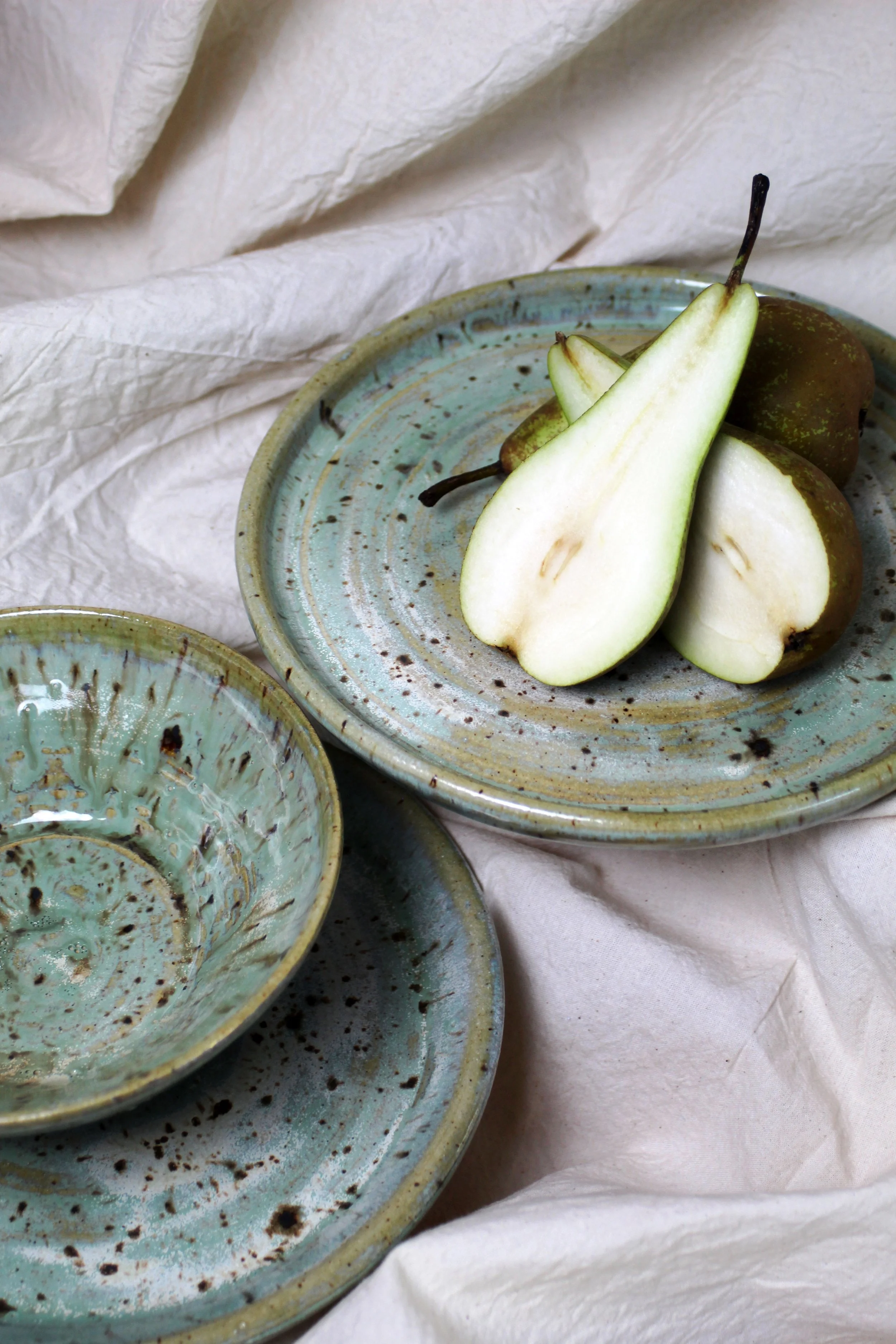 Two rustic teal ceramic plates, one with slices of green pear and whole pears, set on crinkled white fabric.
