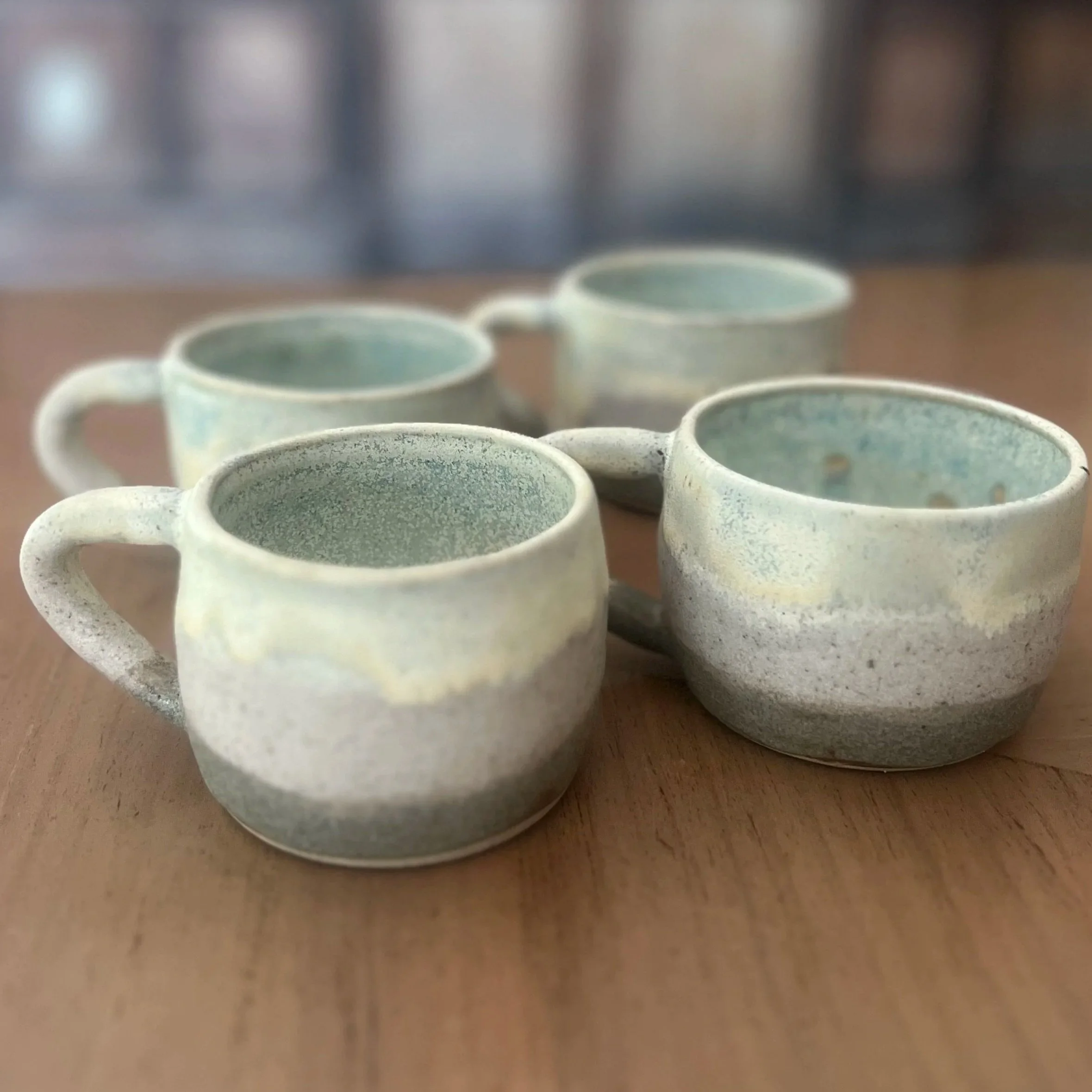 Four handmade stoneware mugs with soft matte white and sandy speckled exteriors and pale celadon blue interior glazes, arranged together on a wooden surface.