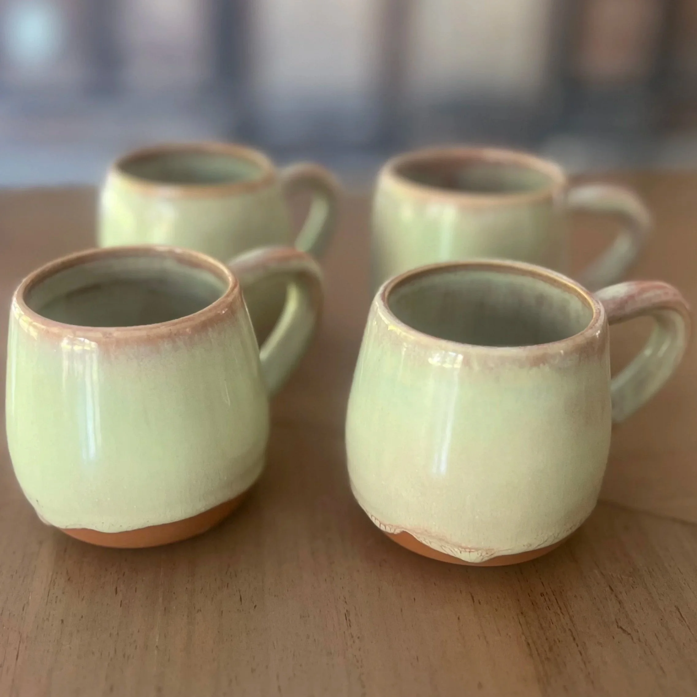 Four handmade terracotta mugs with a glossy soft cream and blush pink glaze and natural unglazed clay base, sitting on a wooden surface.