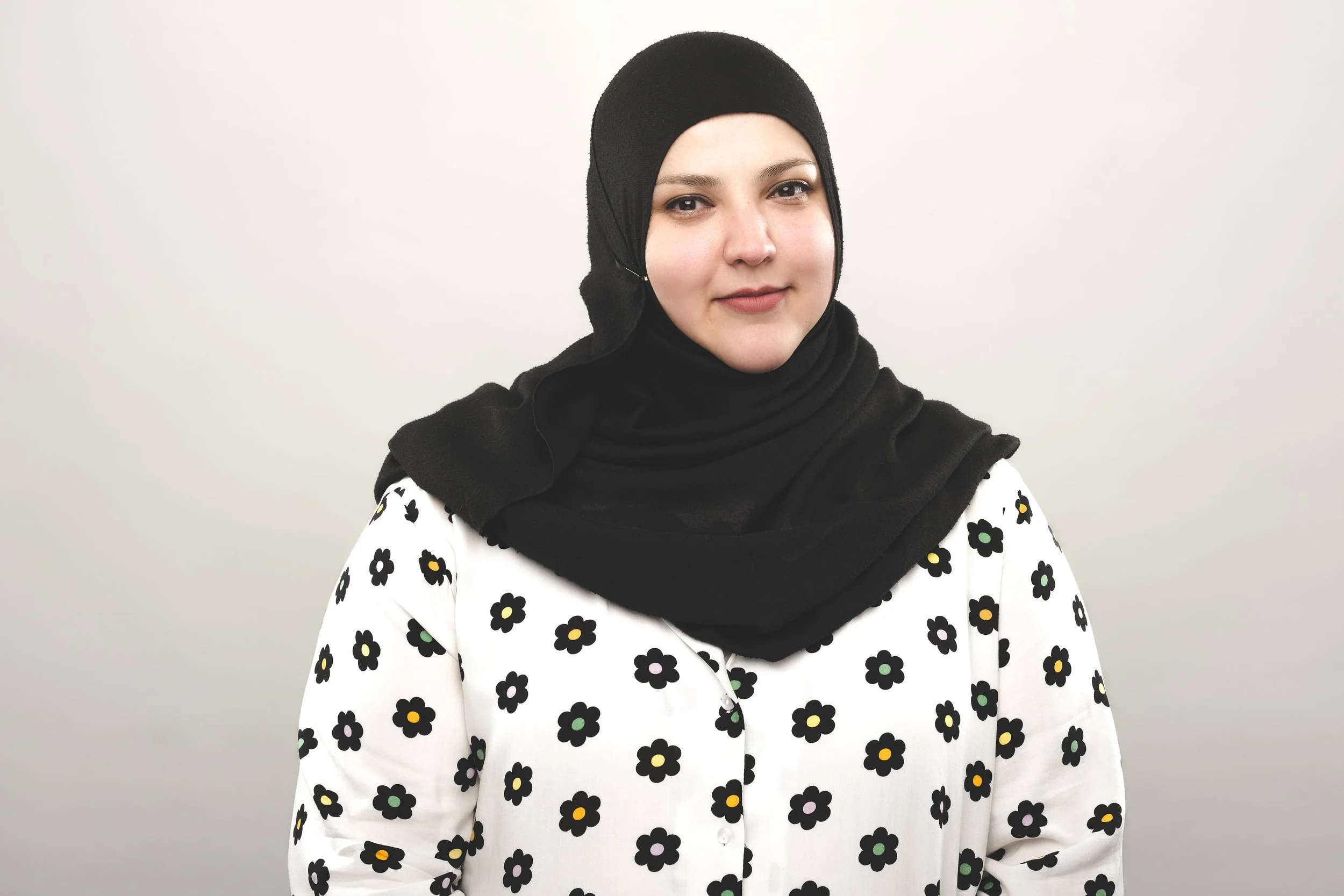 Fatiha El-Ghorri, comedian wearing a black hijab and a white shirt with black and yellow floral pattern against a plain light background.