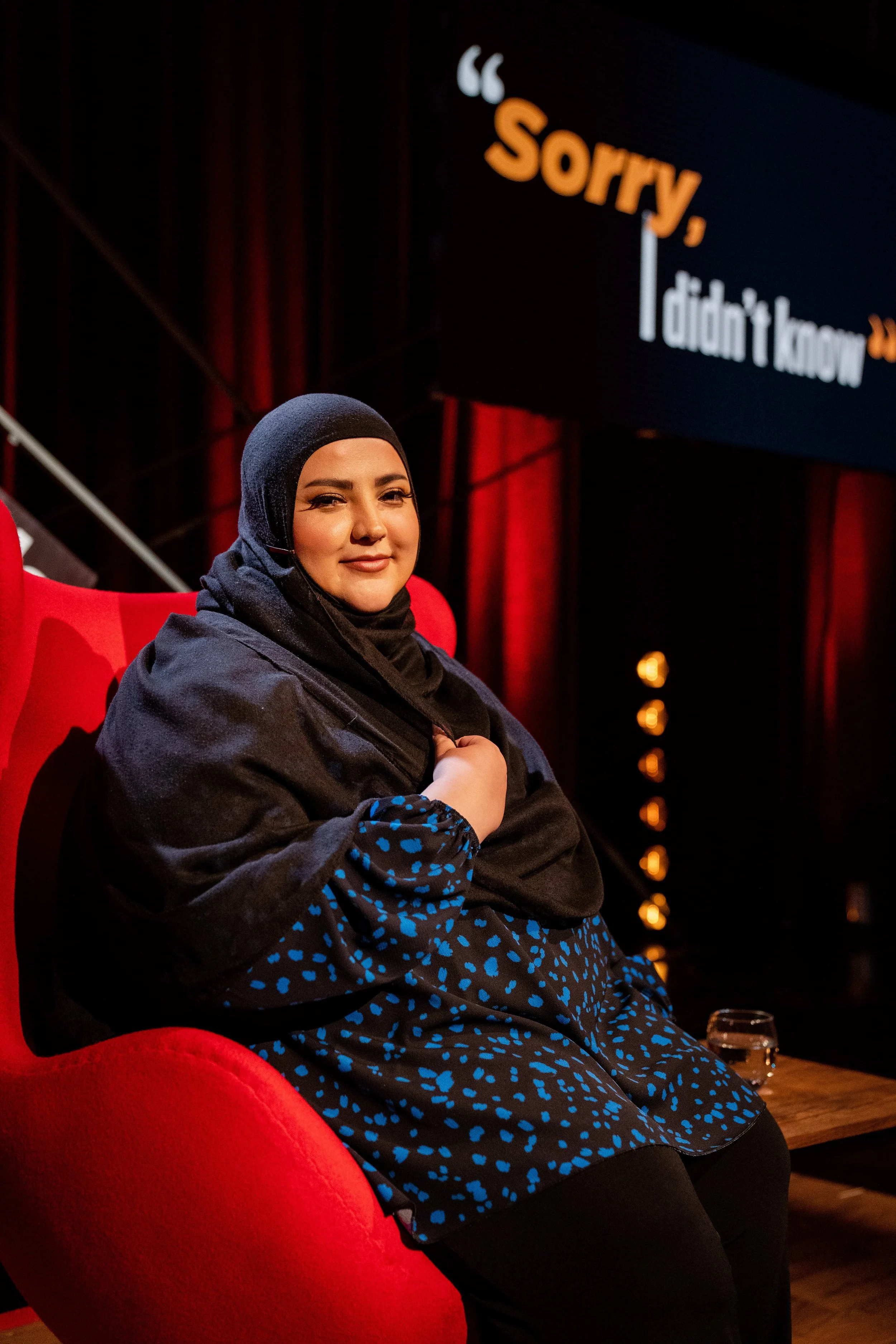 Fatiha El-Ghorri wearing a black hijab and black and blue patterned outfit sitting on a red armchair on stage at a conference or event, with a large screen behind her displaying the phrase 'Sorry, I didn't know' in large text.