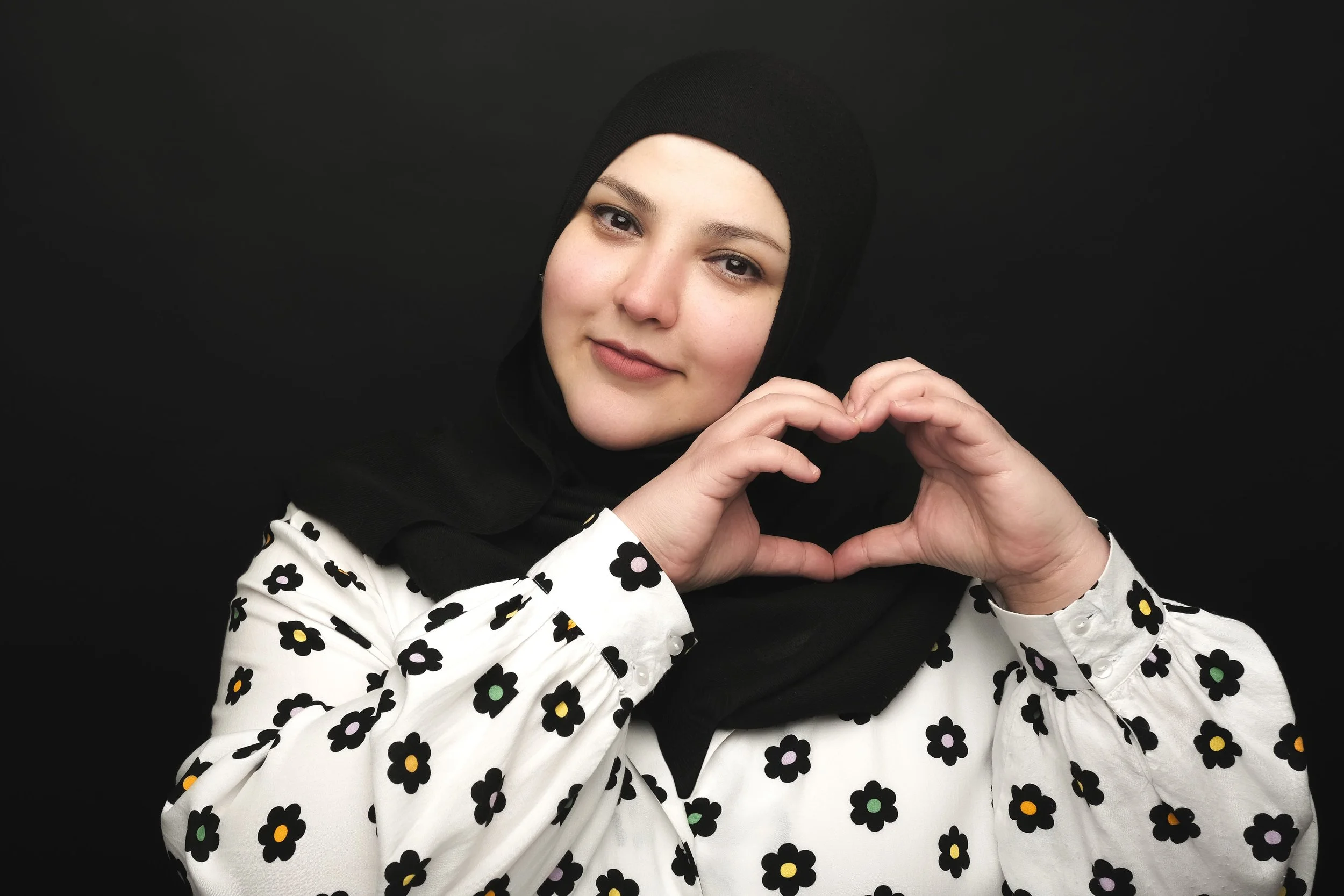 Fatiha El-Ghorri black headscarf and a white blouse with black and yellow flower patterns making a heart shape with her hands, against a black background.