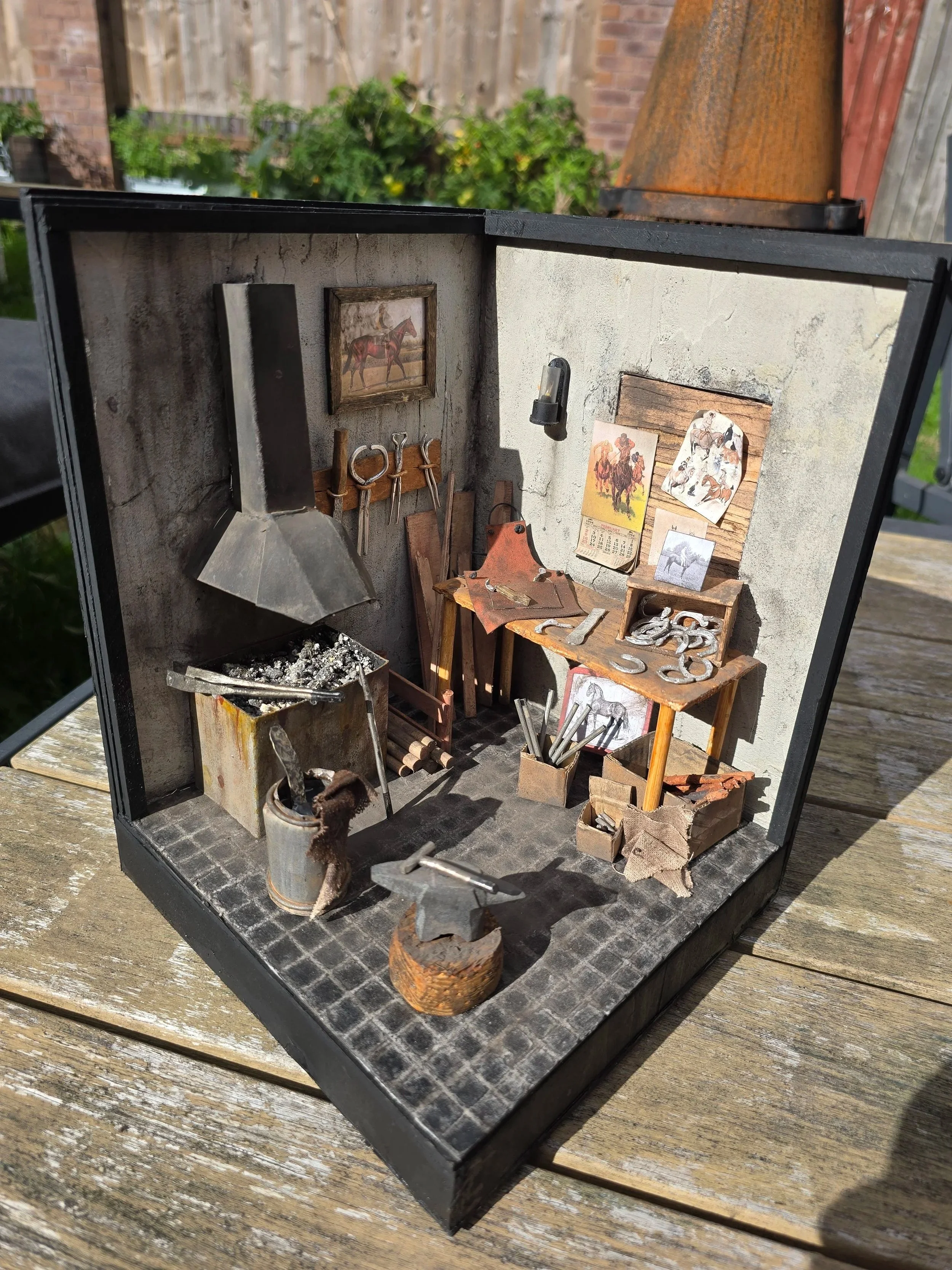 Miniature model of a blacksmith workshop with tools, a small anvil, and pictures of horses on the wall, set on a wooden surface outdoors.