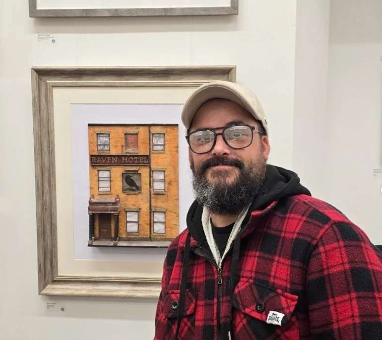 A man with glasses, a beard, wearing a baseball cap and red plaid jacket, standing in front of a framed painting of a building with a sign that reads 'Raven Hotel' and a black raven image.