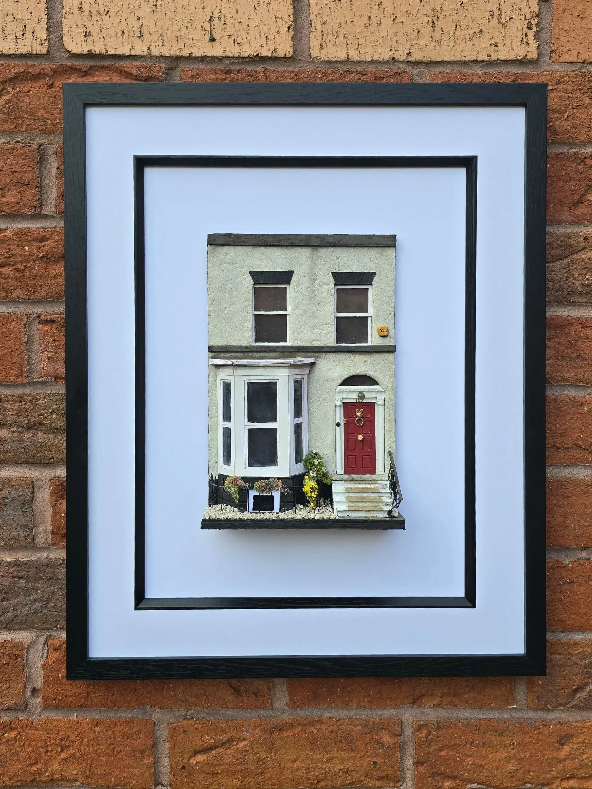 A framed photograph of a house with a green exterior, red door, and front steps, hanging on a brick wall.