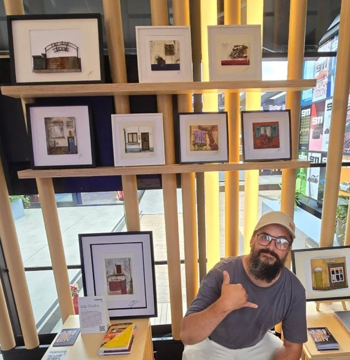 Man with glasses and a beard making a shaka sign while sitting in front of framed artwork displayed on wooden vertical slats in a gallery or store.