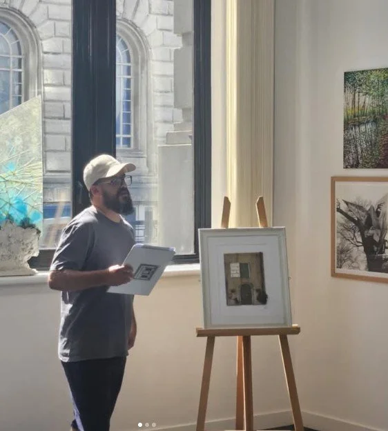 Man in a gray shirt and white baseball cap viewing artwork displayed on an easel inside an art gallery.