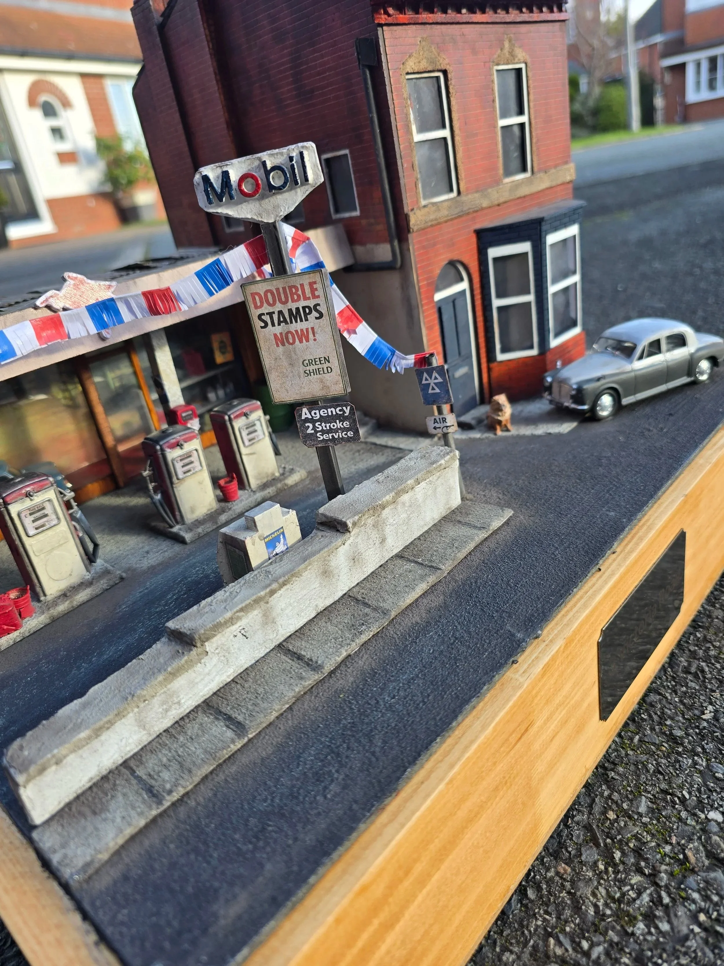 A miniature model of a gas station with vintage gas pumps, a small building with a storefront, and a tall brick residential building in the background. A gray toy car is parked on the street.