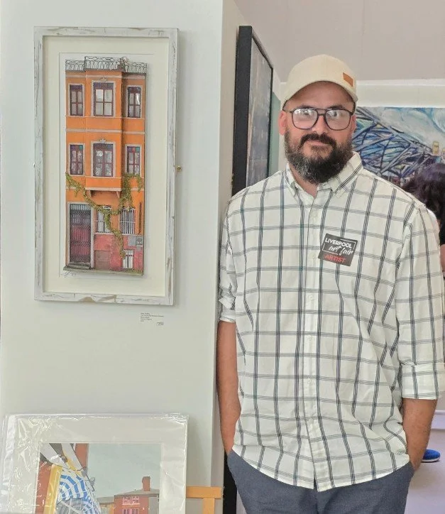 A man with a beard, glasses, and a beige cap standing indoors near artwork on the wall, wearing a plaid shirt with a "Liverpool" sticker, and hands in pockets.