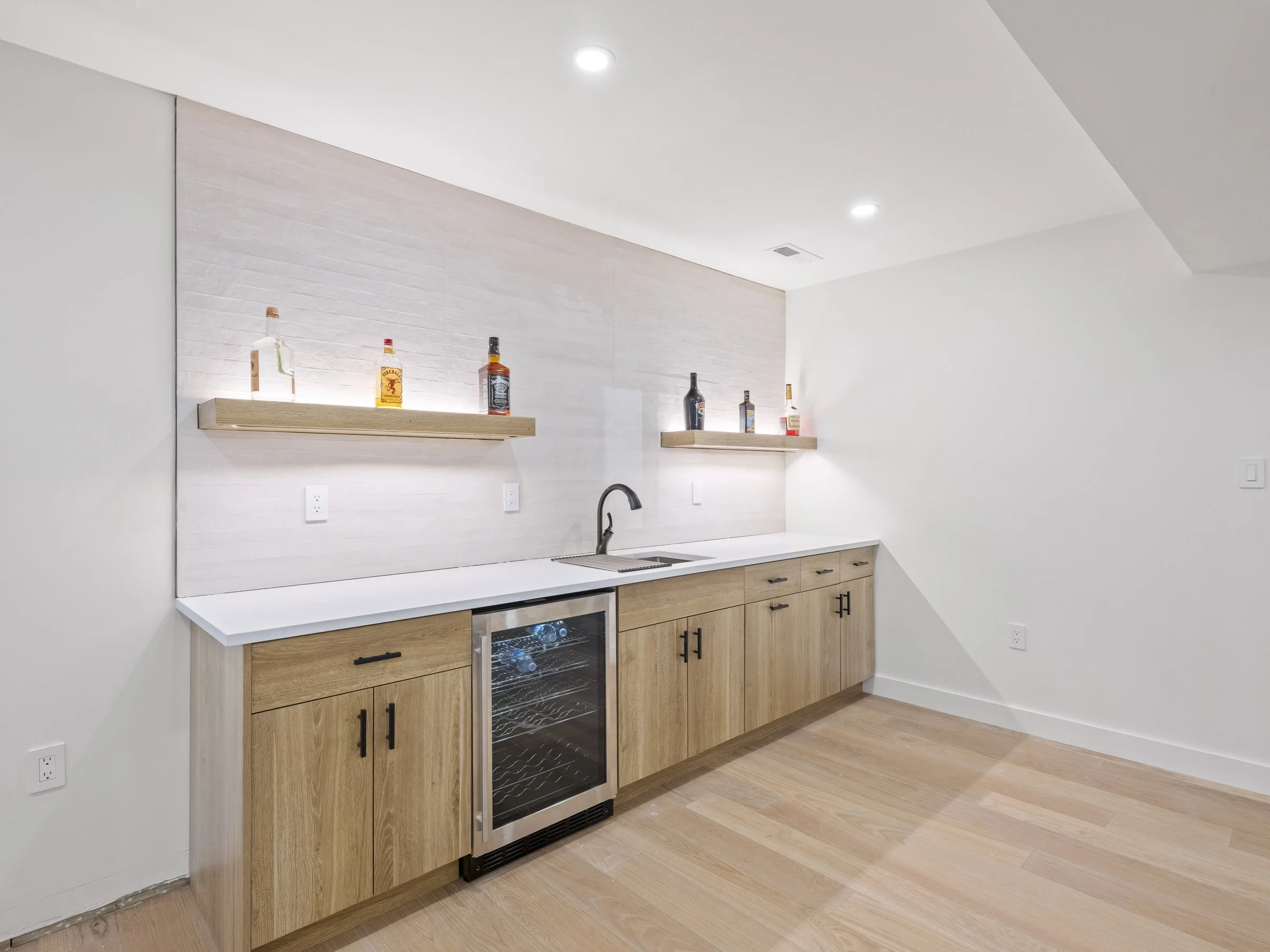 Modern kitchenette with wooden cabinets, a white countertop, and a mini wine fridge. Two floating shelves display various liquor bottles. The space has light-colored walls and flooring with recessed ceiling lights.