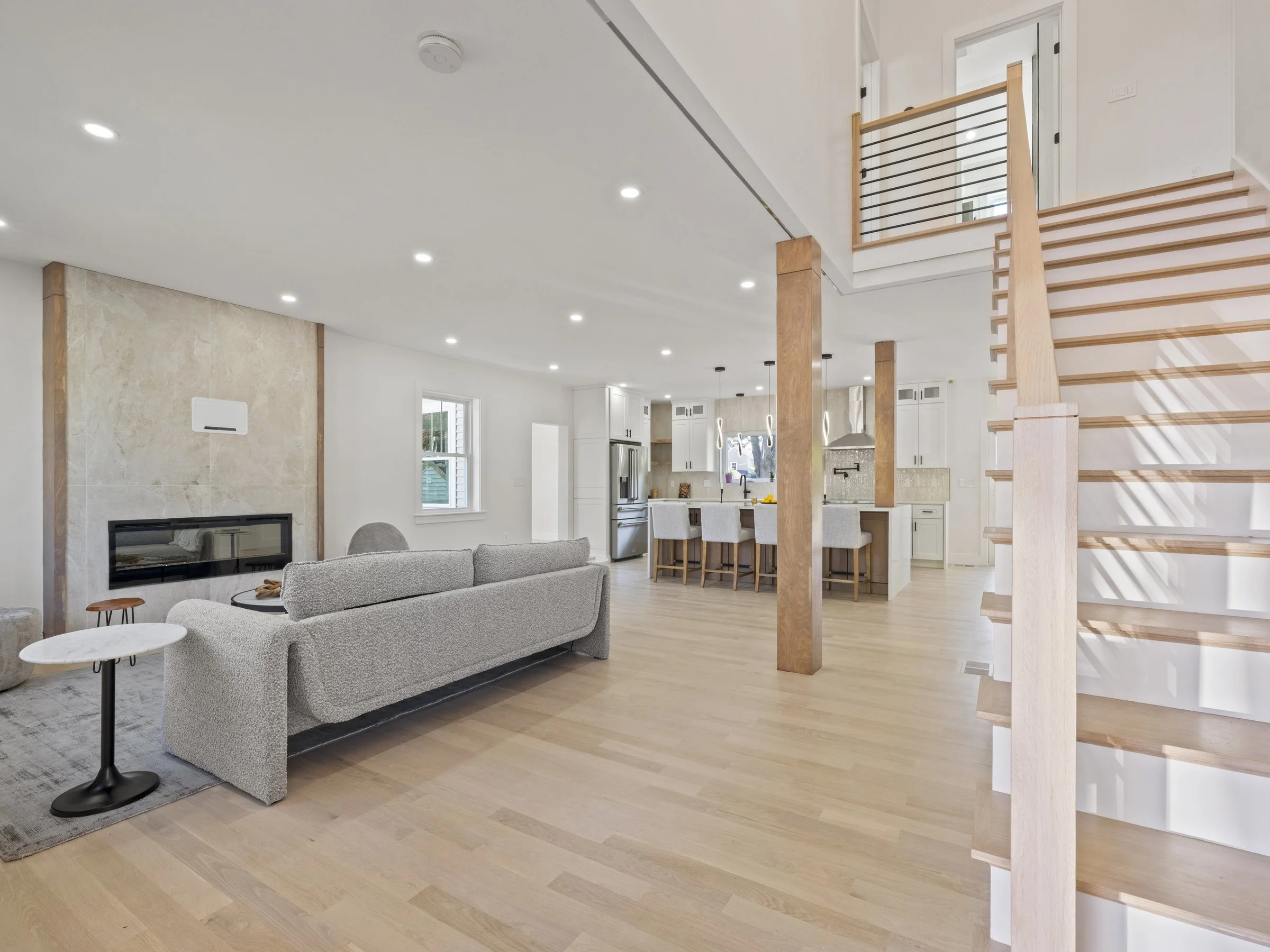 Open-concept living room and kitchen with light wood flooring, white walls, modern furniture, staircase, and large windows.
