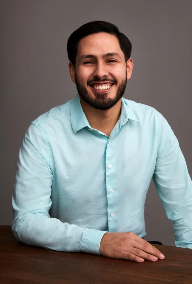 A man with dark hair and a beard, wearing a light blue button-up shirt, smiling, seated at a dark wooden table against a gray background.