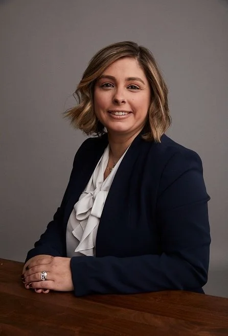 A professional woman with blonde hair in a bob style, wearing a dark blazer and a white blouse with a bow, sitting at a wooden table against a gray background.